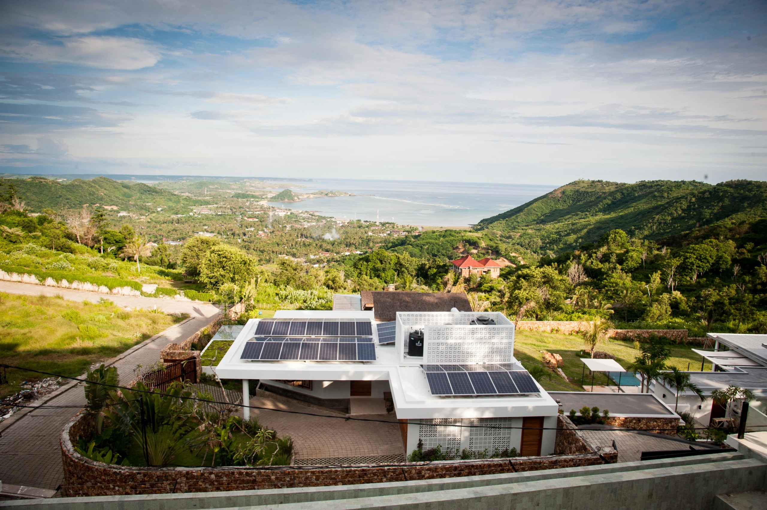 High-angle view of a coastal villa roof equipped with solar panels overlooking hills and the ocean.