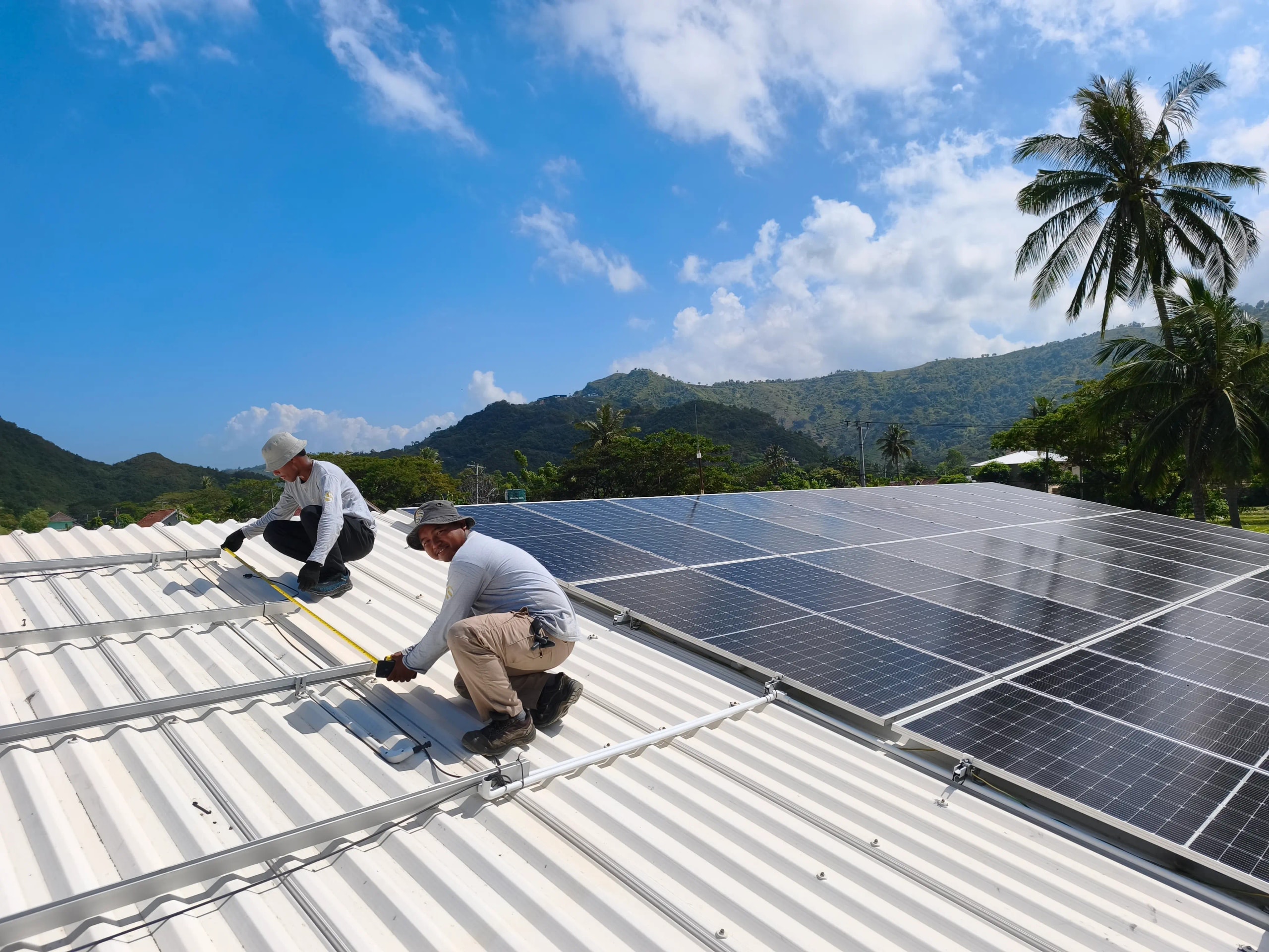 Two technicians installing solar panel frames on a light-colored corrugated rooftop against a mountain backdrop.