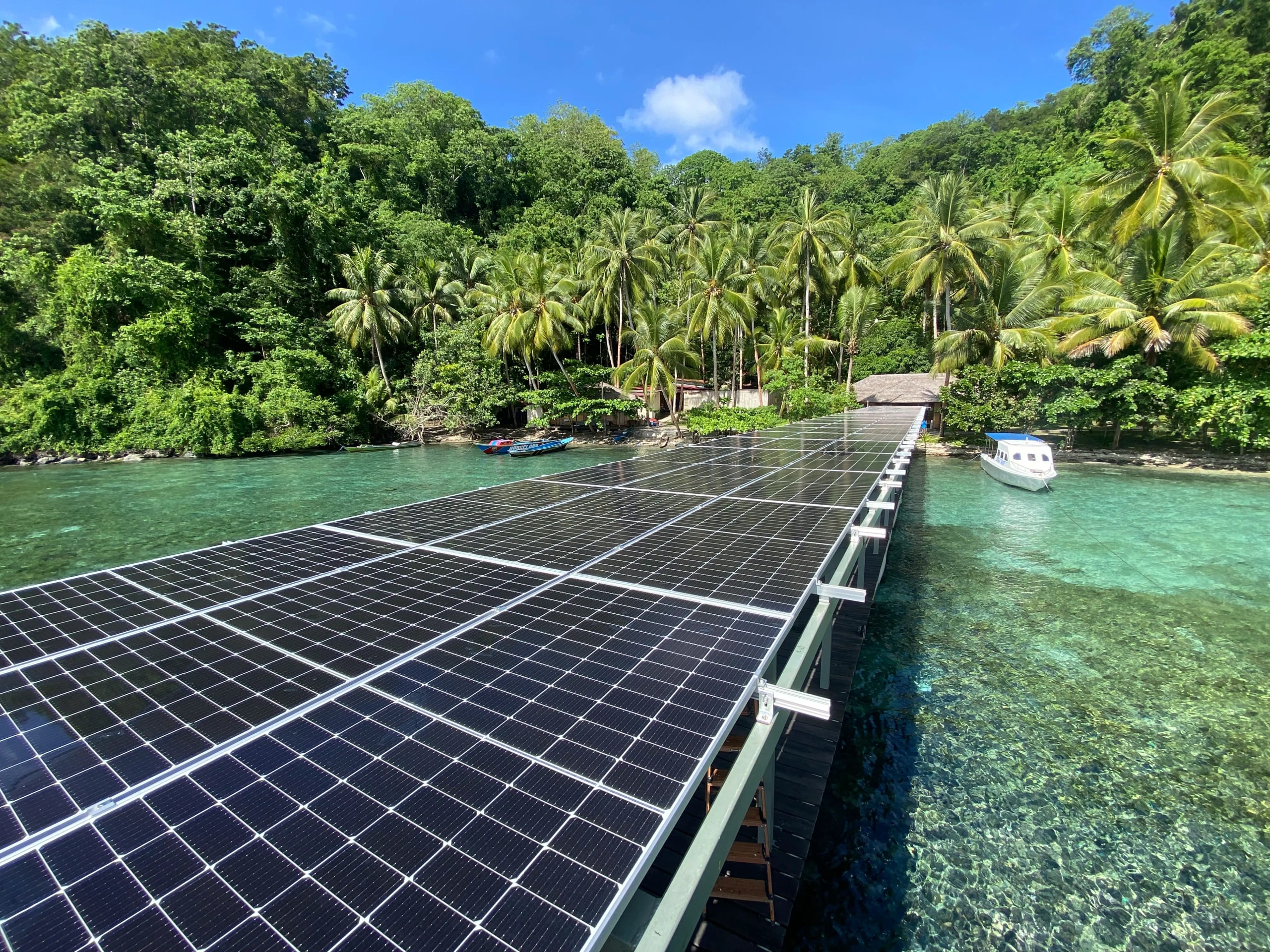 A long wooden pier lined with solar panels extending into clear turquoise ocean waters near a tropical shore.