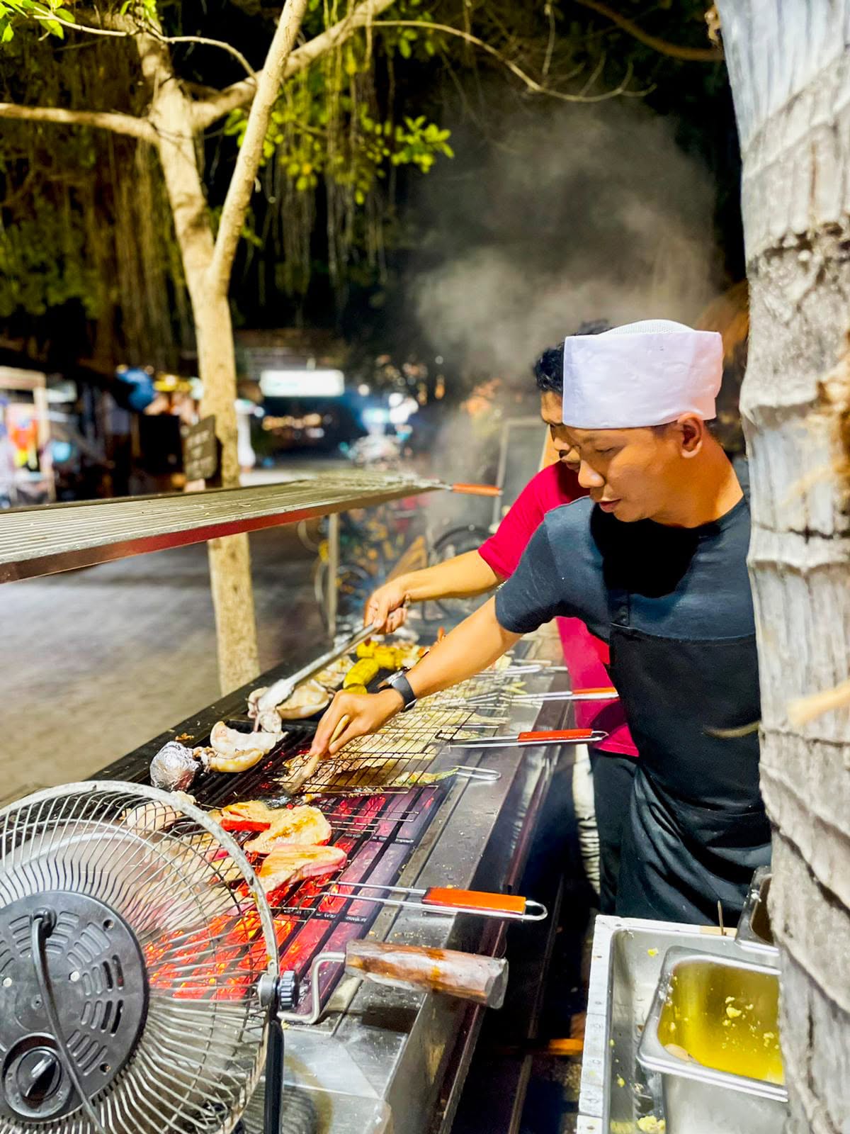 Chef in a white hat grilling seafood and meat skewers over an open flame barbecue.