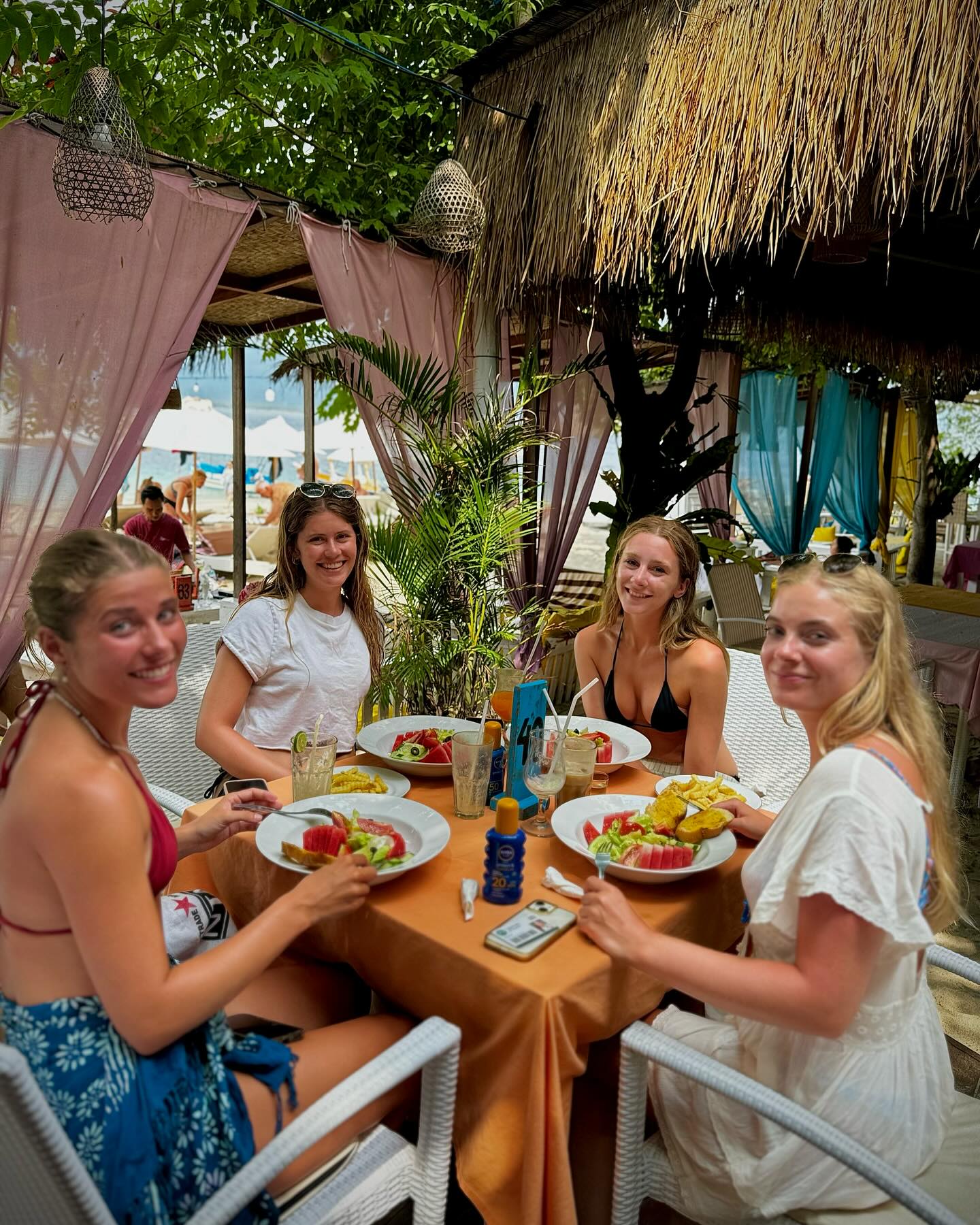 Four women smiling at a dining table under a thatched roof structure with pink curtains.
