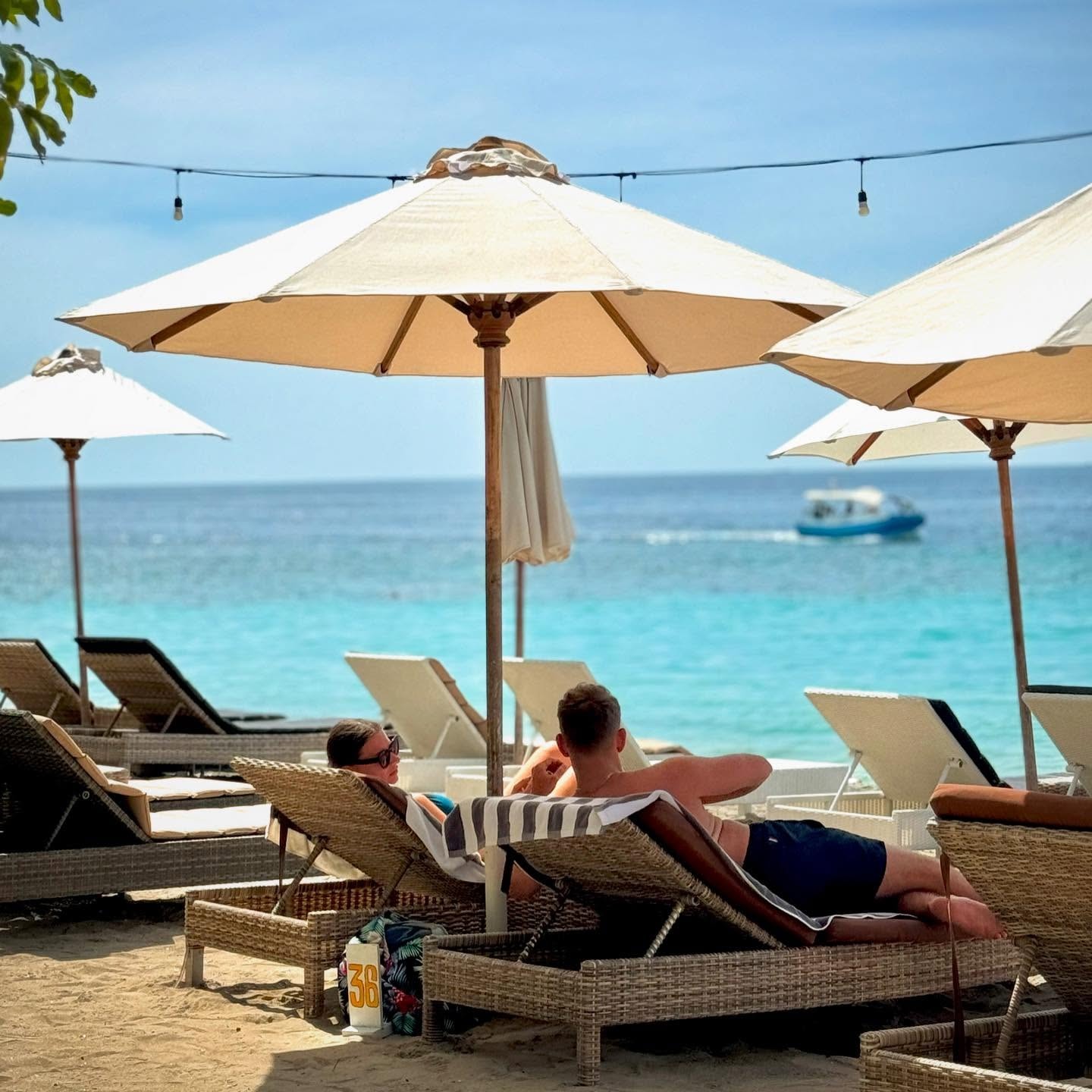 People relaxing on woven beach sun loungers under white umbrellas looking out at the turquoise sea.