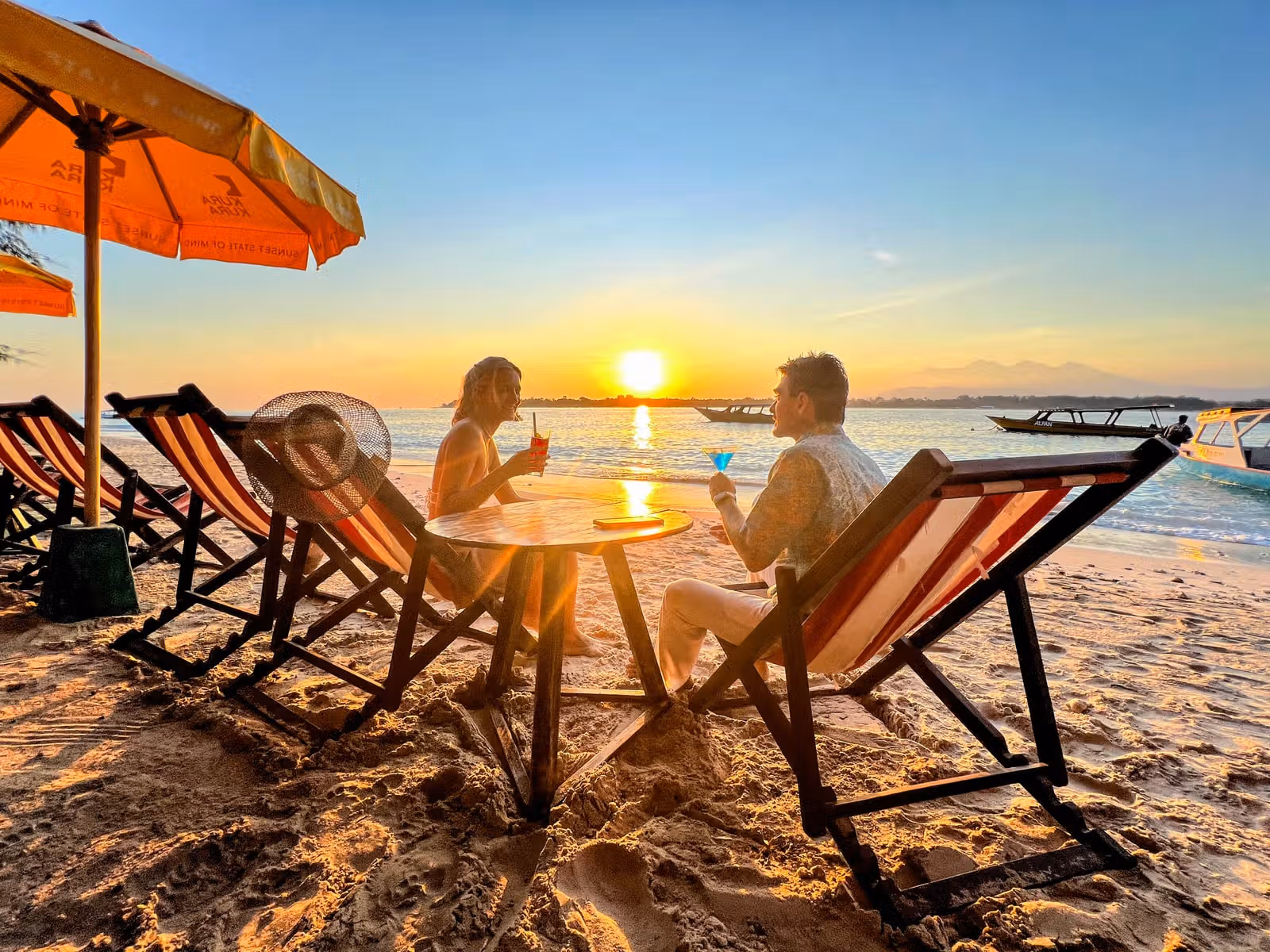 A couple enjoying cocktails in lounge chairs on the beach while watching the sunset.