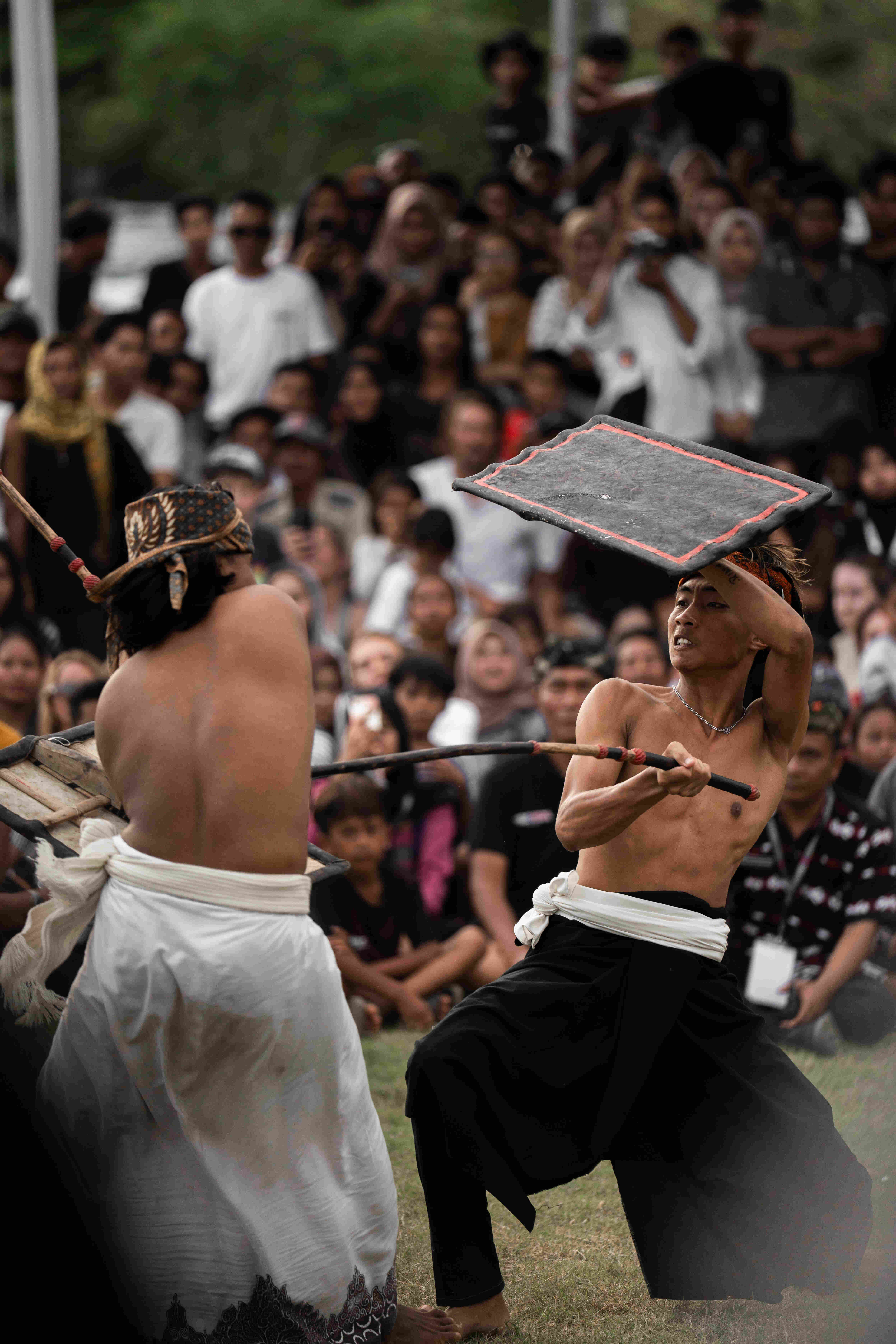 Live-action view of a dynamic Peresean (Sasak stick fighting) duel in Lombok, showing two pepadu fighters mid-action with traditional sticks and shields, surrounded by an engaged crowd of spectators.