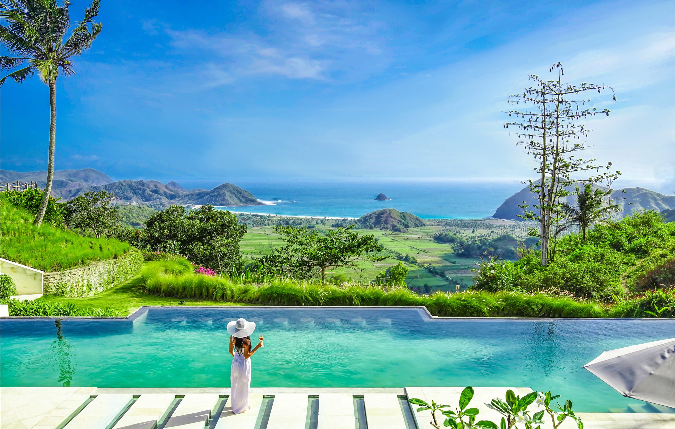 Woman in a white dress standing by an infinity pool overlooking a lush landscape and ocean view under a clear blue sky.