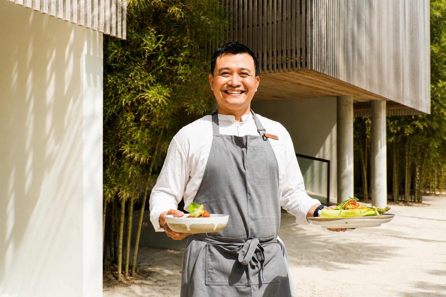 Chef Matthew Angga smiling while holding two gourmet plates of Indonesian food outside a resort building.