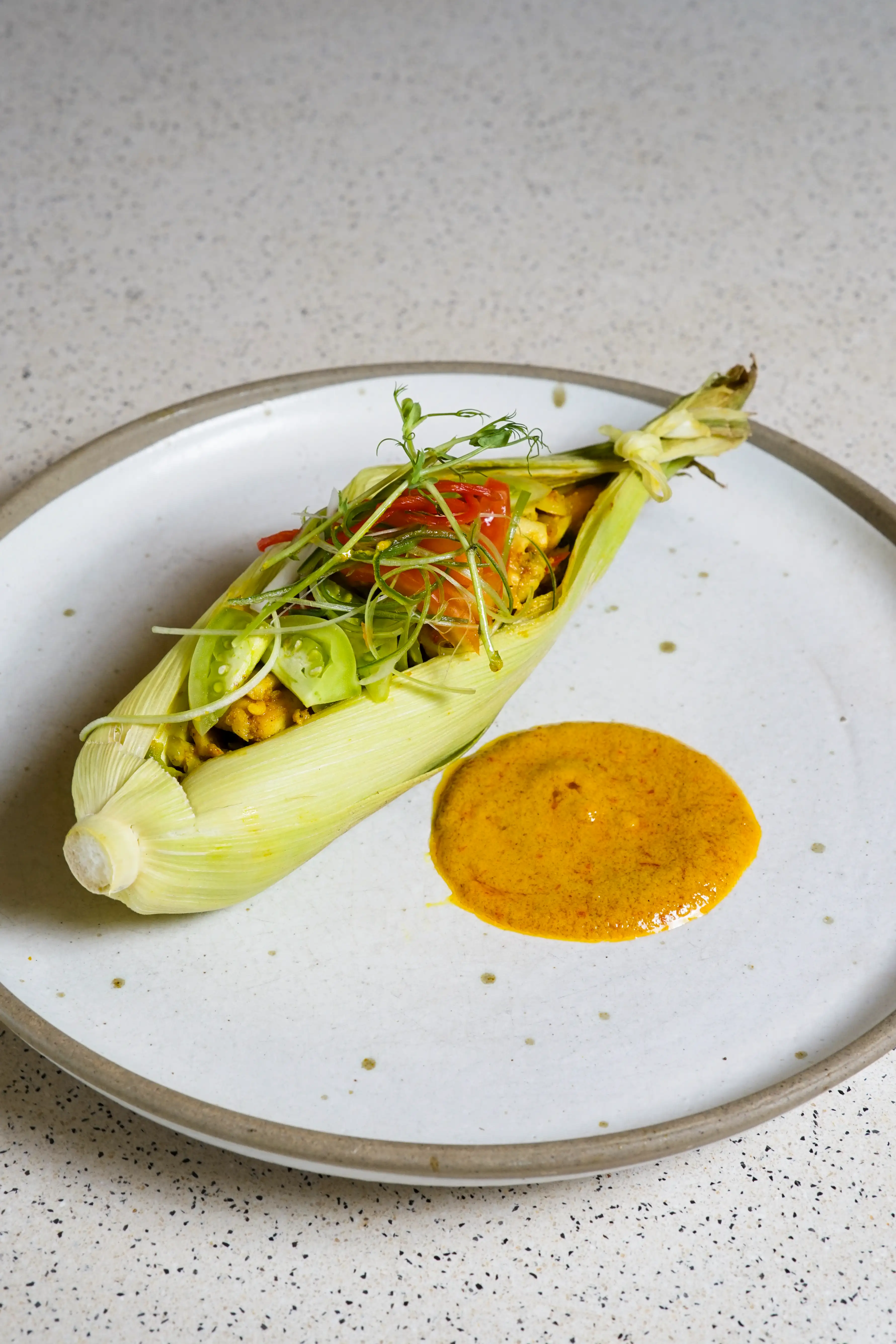 Garnished corn-husk wrap next to orange sauce on a speckled plate.