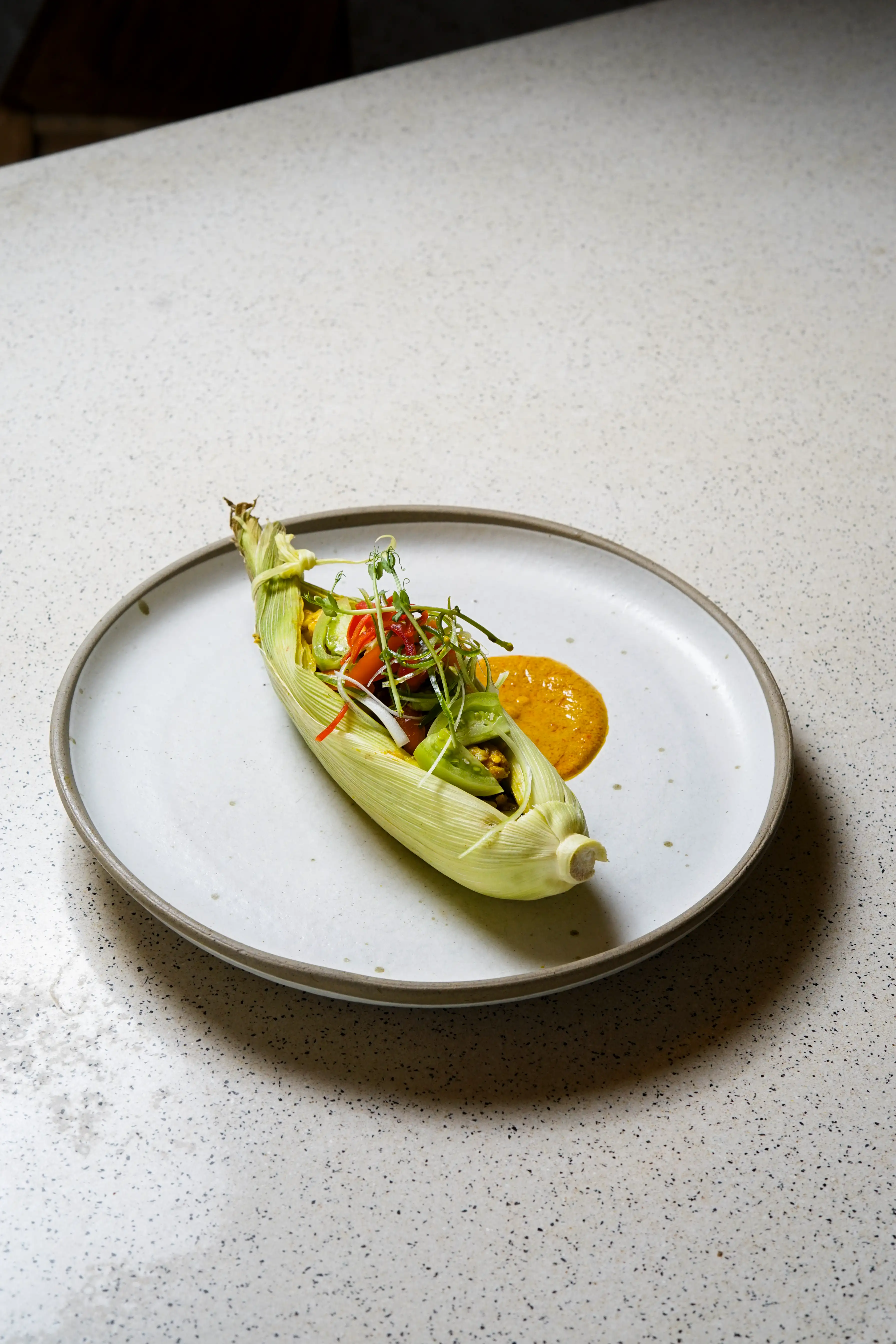 Modern Pepes Ikan served in a corn husk wrapper with fresh garnishes and spice paste on a white plate.