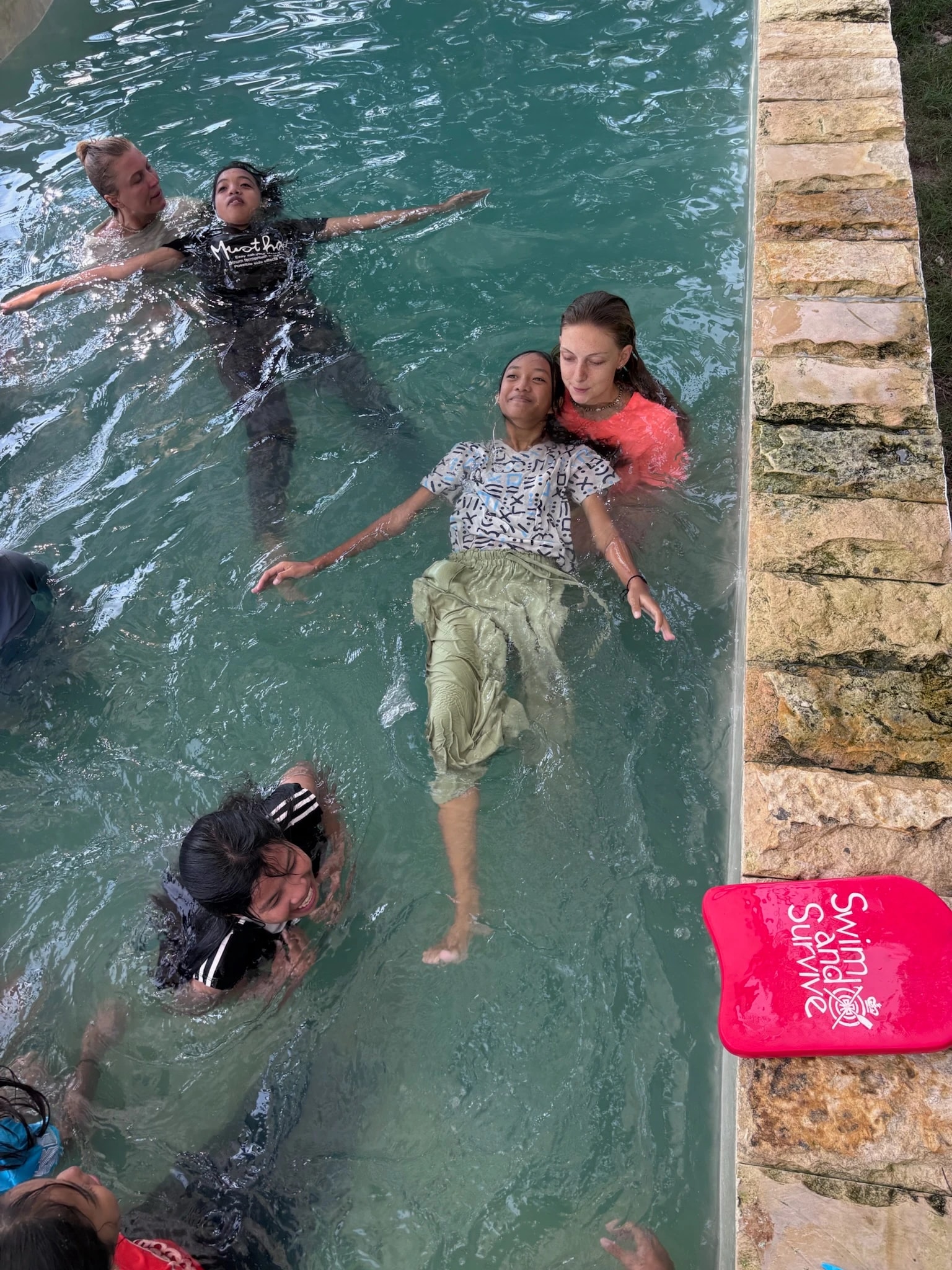 Instructors assisting young girls as they practice floating on their backs in an outdoor swimming pool.