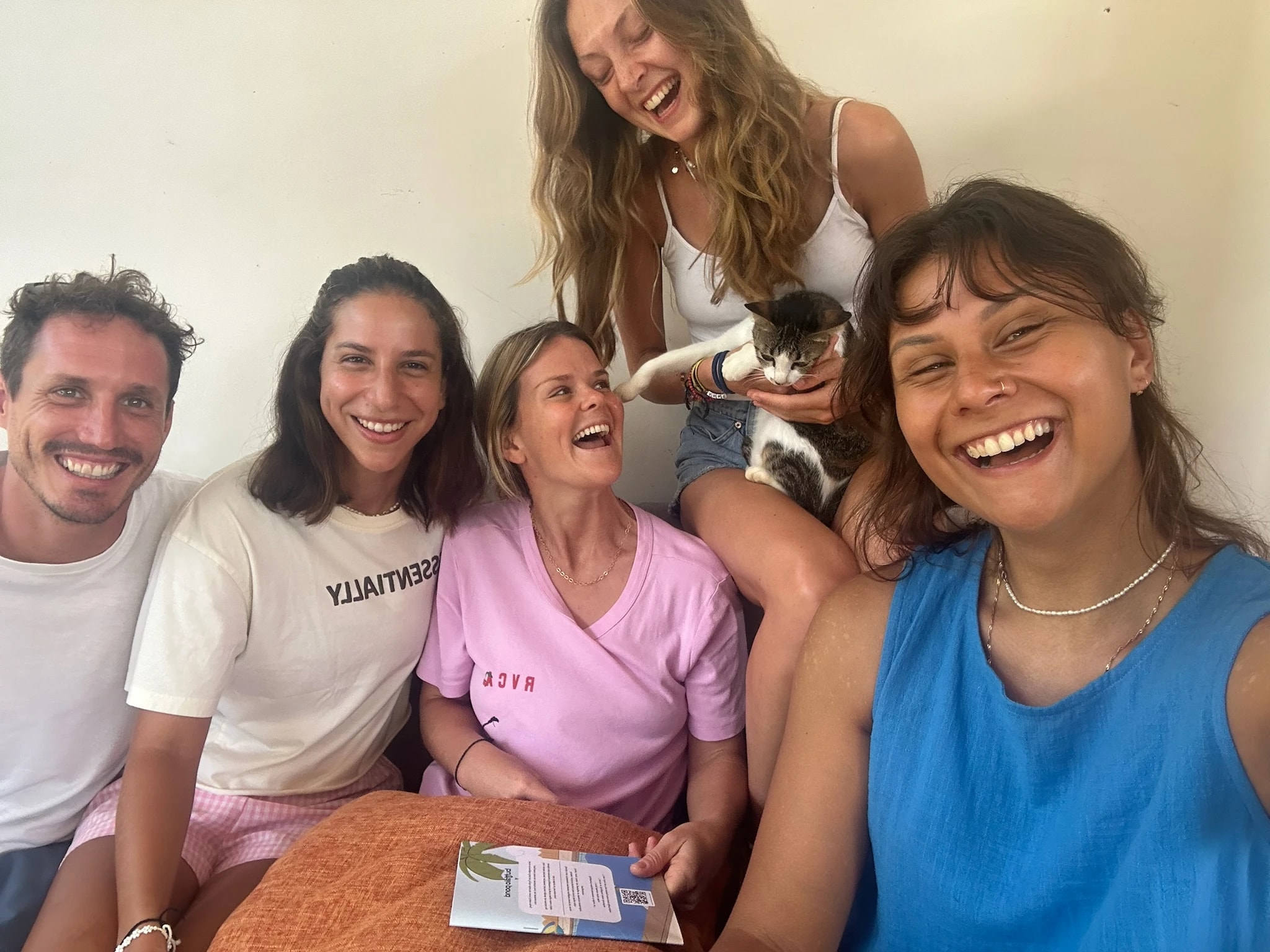 Group of friends smiling and laughing together while taking a close-up selfie indoors with a cat.