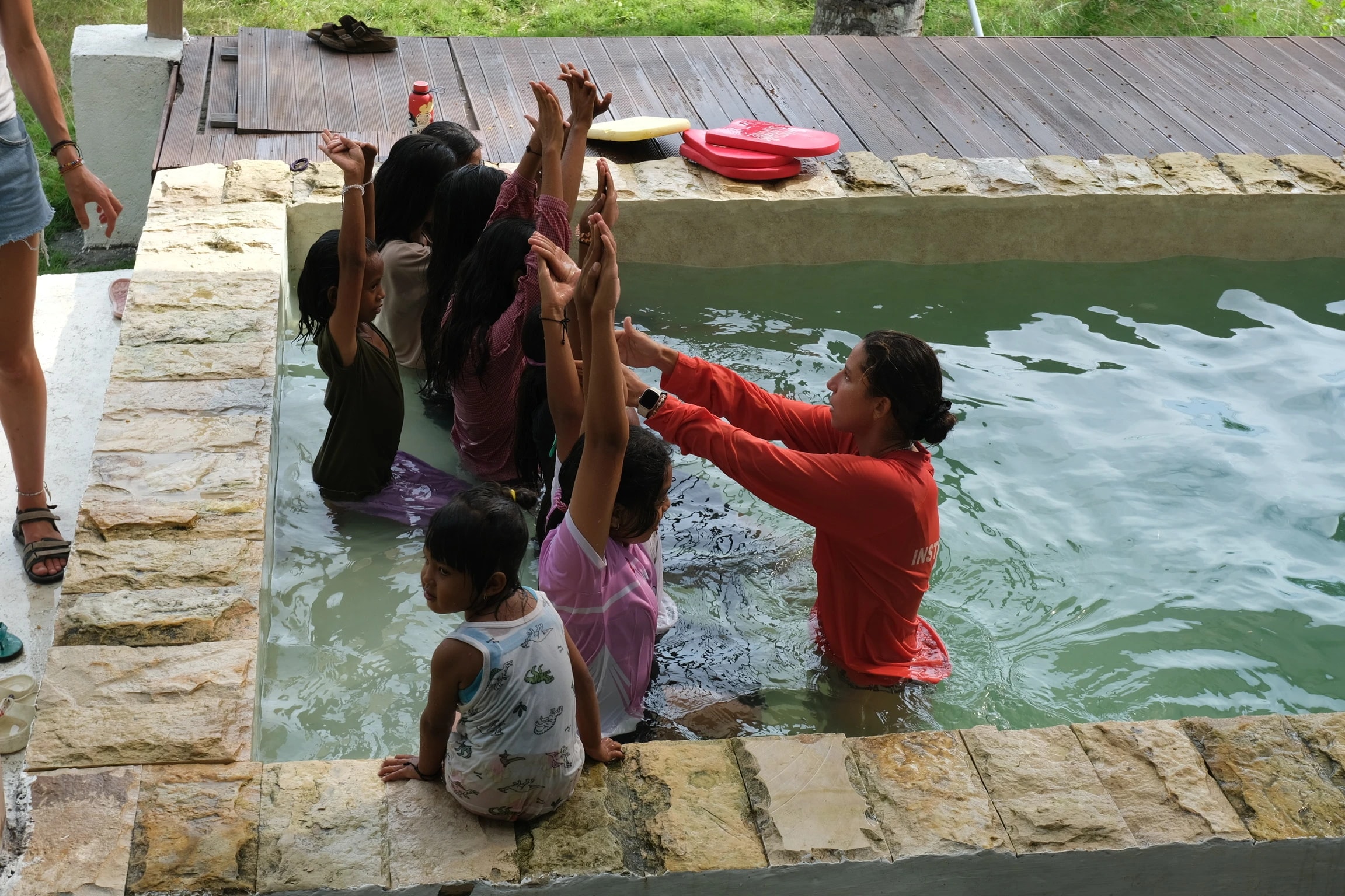 Swim instructor teaching children streamline body position with raised arms along the edge of a pool.