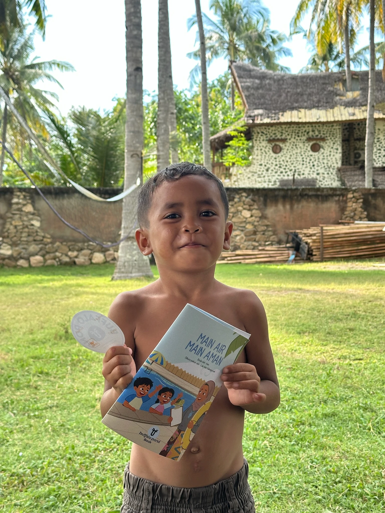Young boy standing outdoors in a yard holding a water safety booklet and a sticker while smiling.