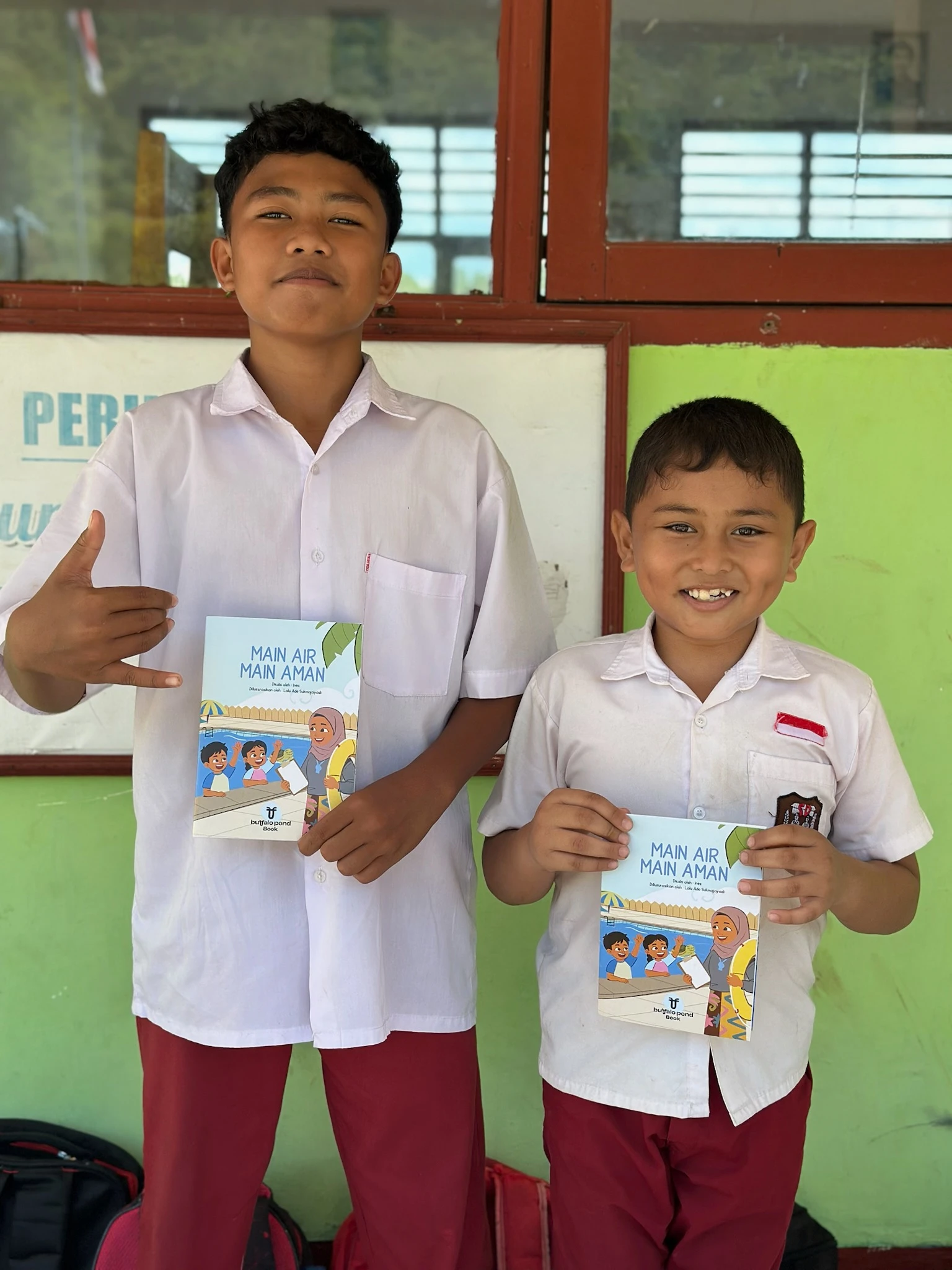 Two schoolboys in Indonesian uniforms proudly holding up water safety booklets in front of a classroom building.