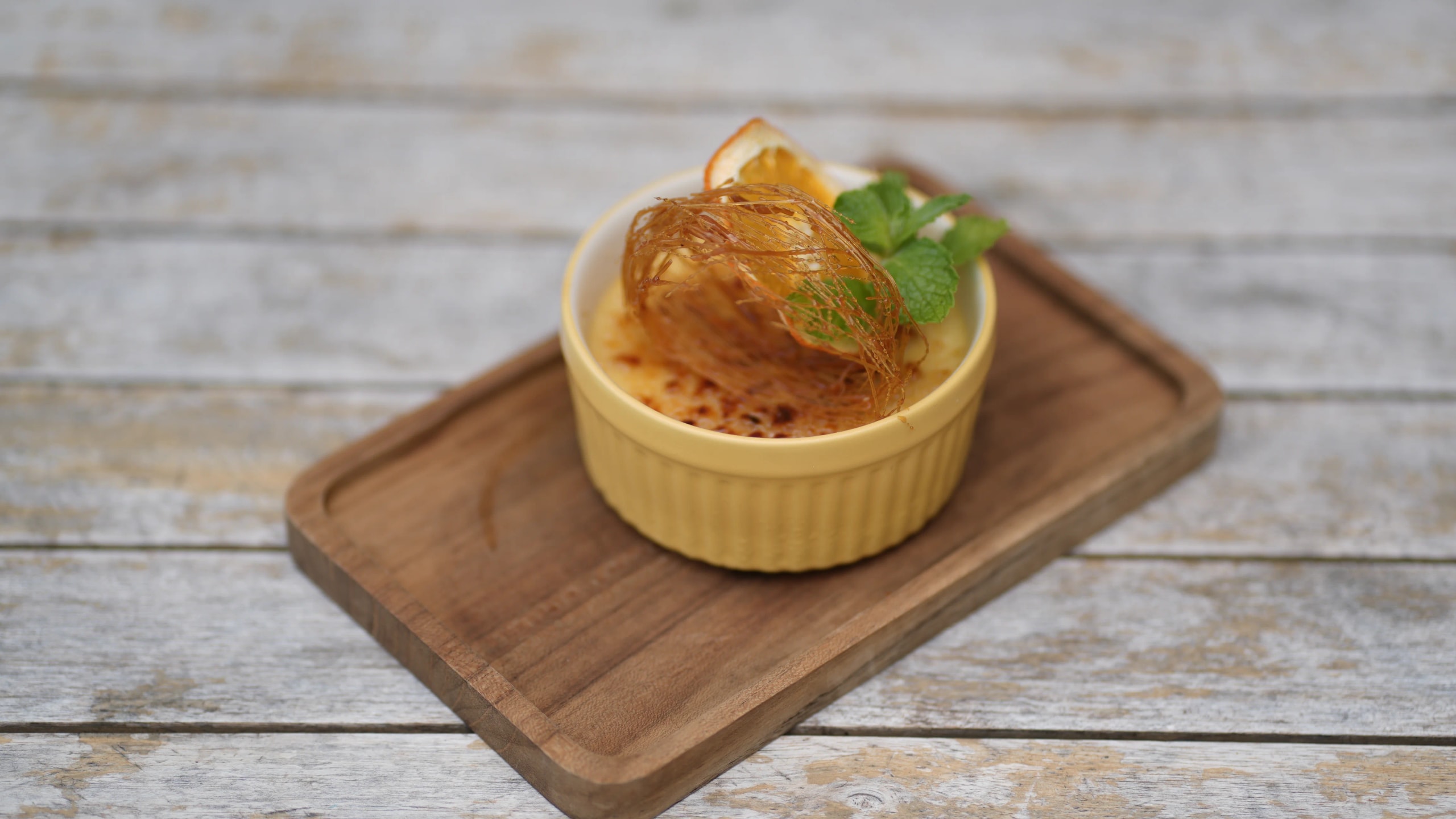 Crème brûlée in a yellow ramekin garnished with a dried citrus slice and spun sugar, served on a wooden tray.