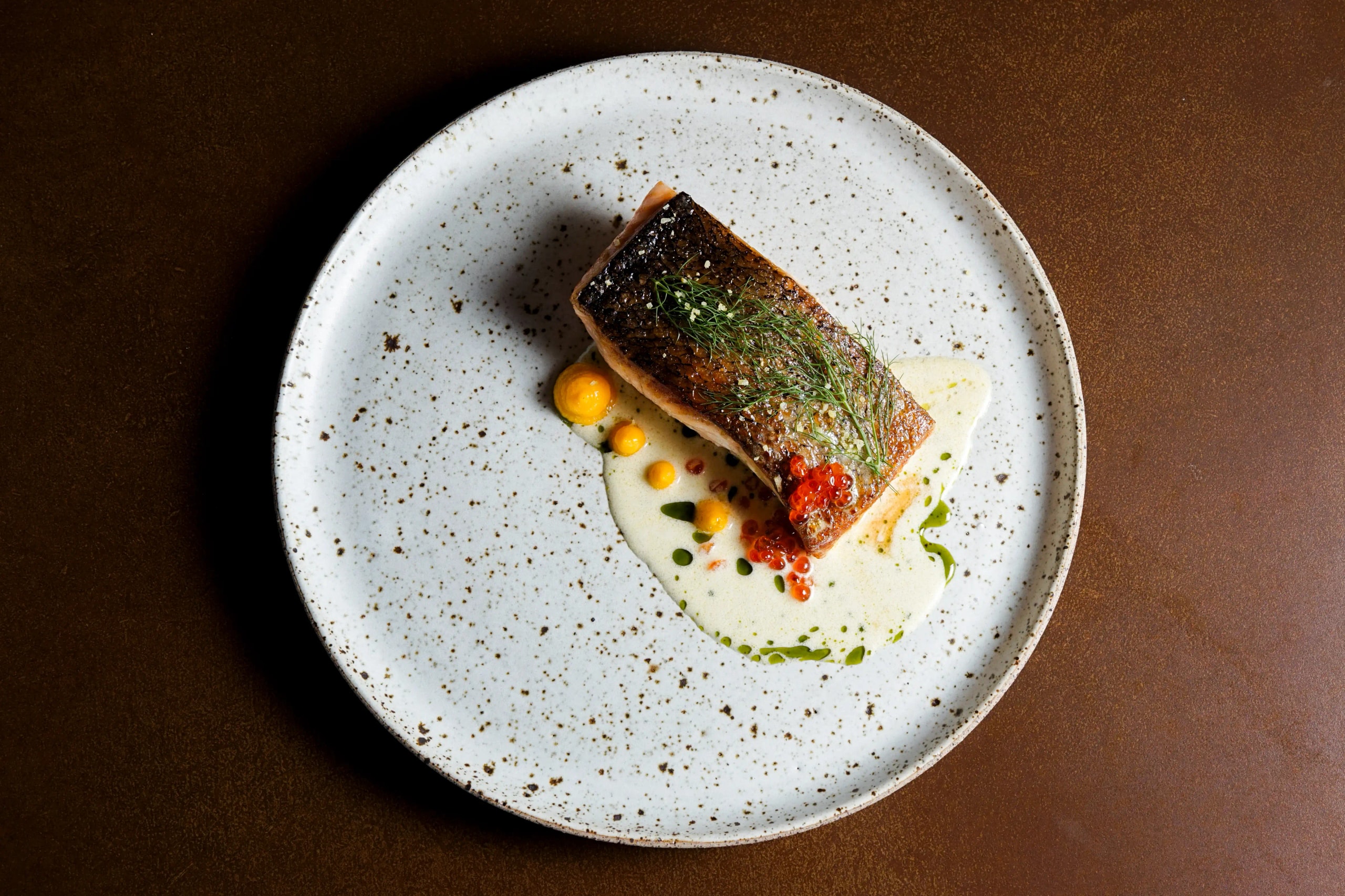 Top-down view of pan-seared fish with salmon roe, herbs, and yellow purée dots in creamy sauce.