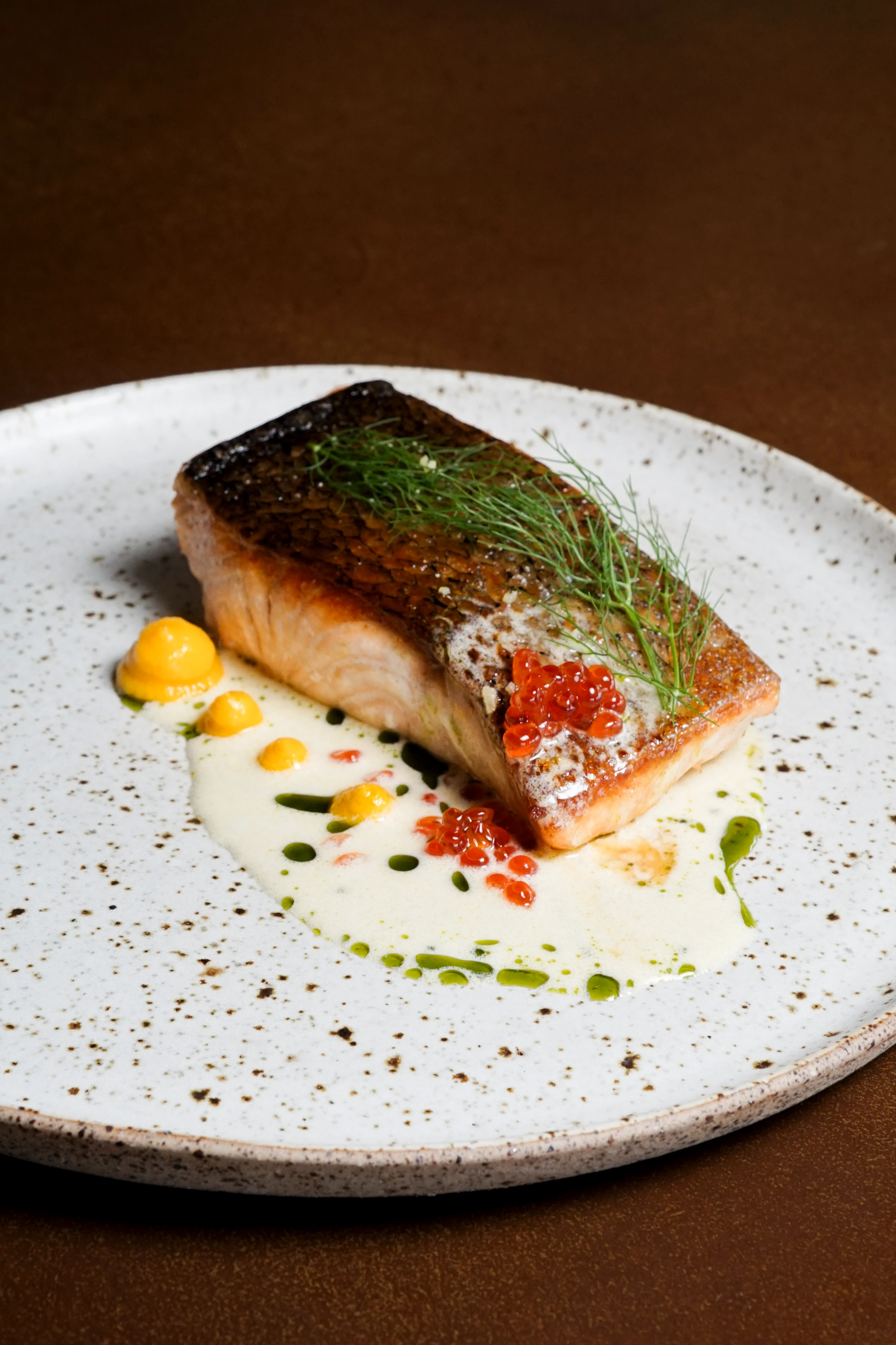 Pan-seared fish fillet topped with salmon roe and dill, served with beurre blanc sauce on a speckled plate.