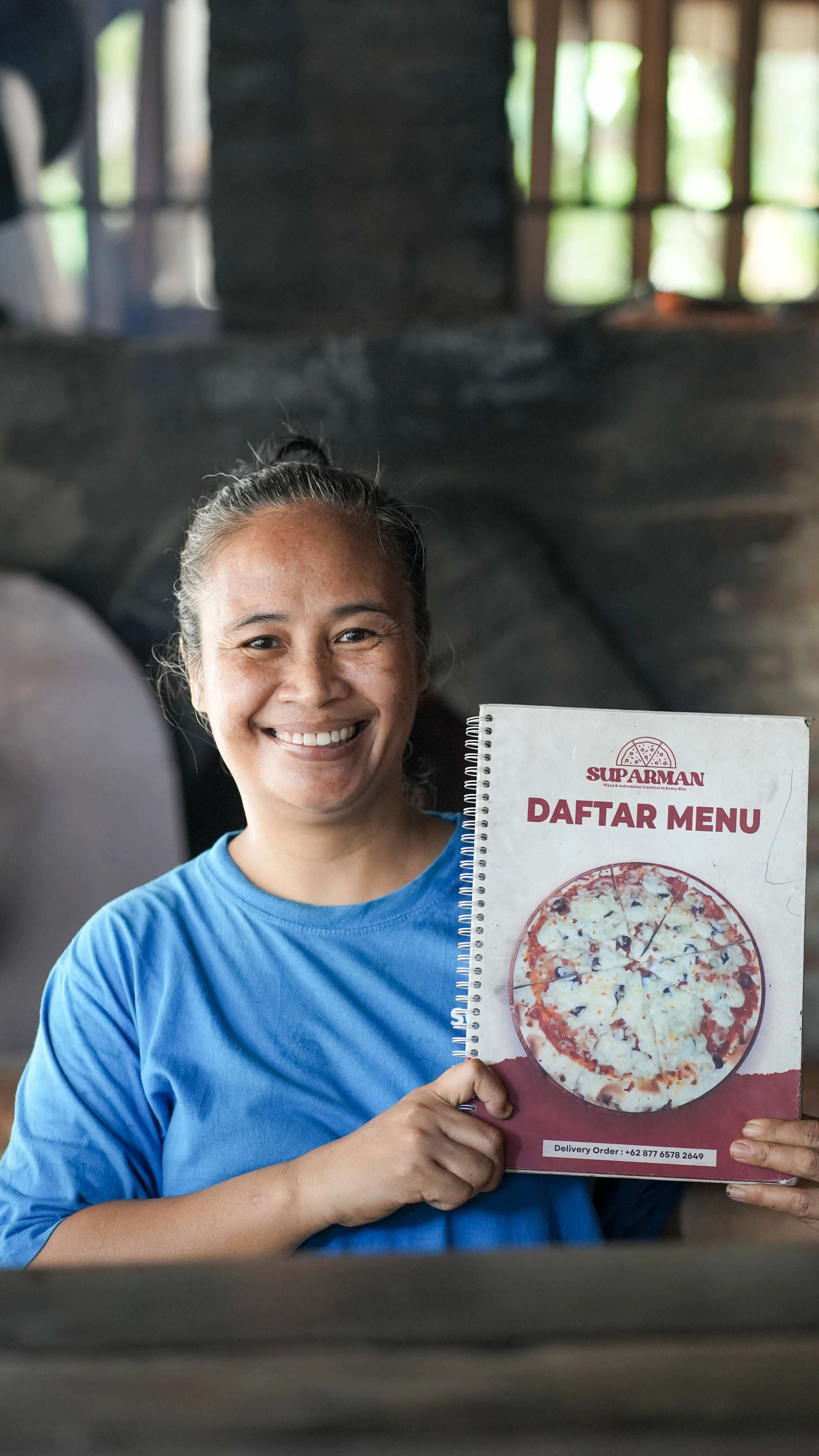 A smiling woman holding up a spiral-bound menu book titled "Suparman Daftar Menu"