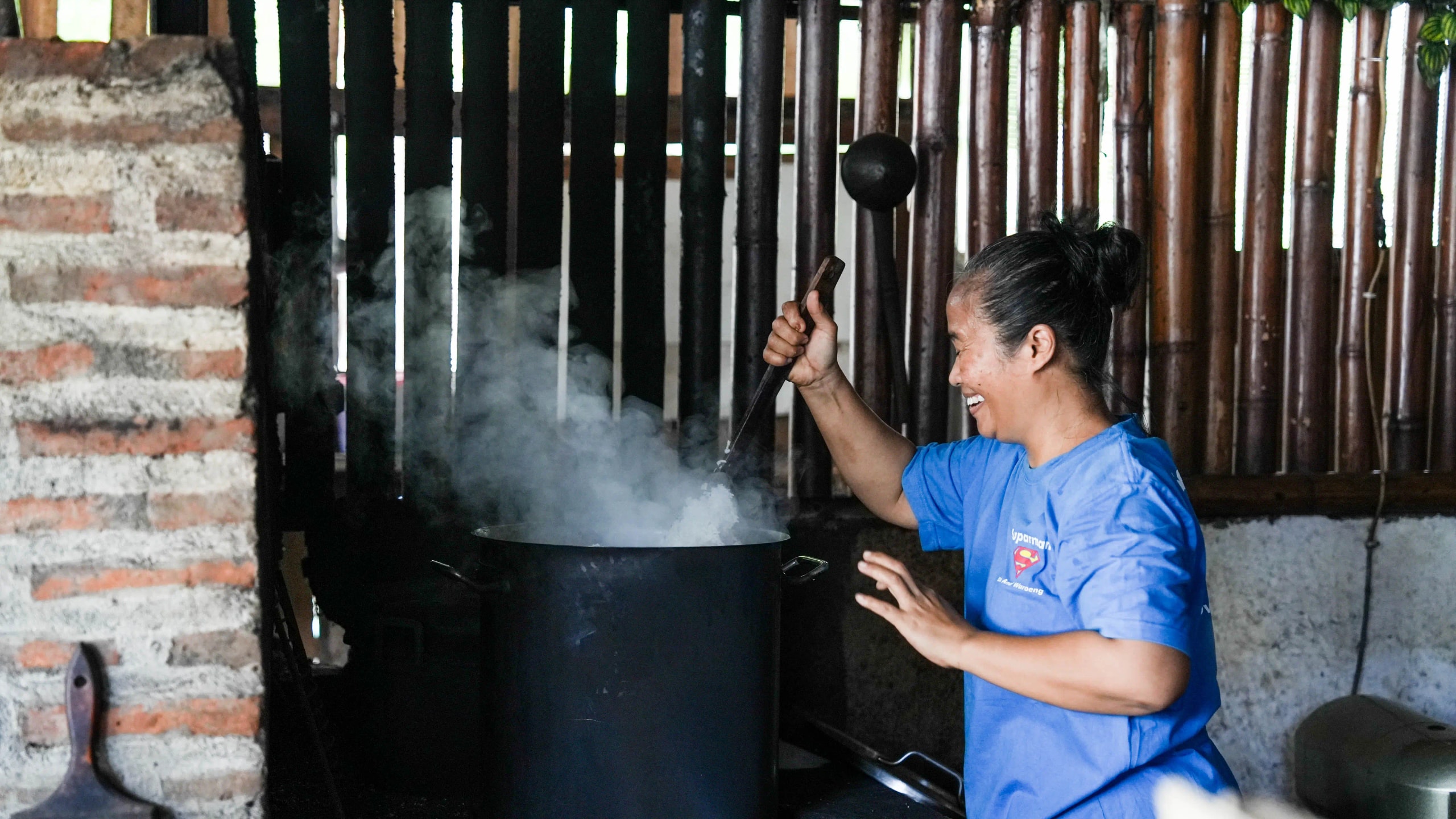 A woman smiling while stirring a steaming pot of food over a traditional stove in a rustic kitchen.