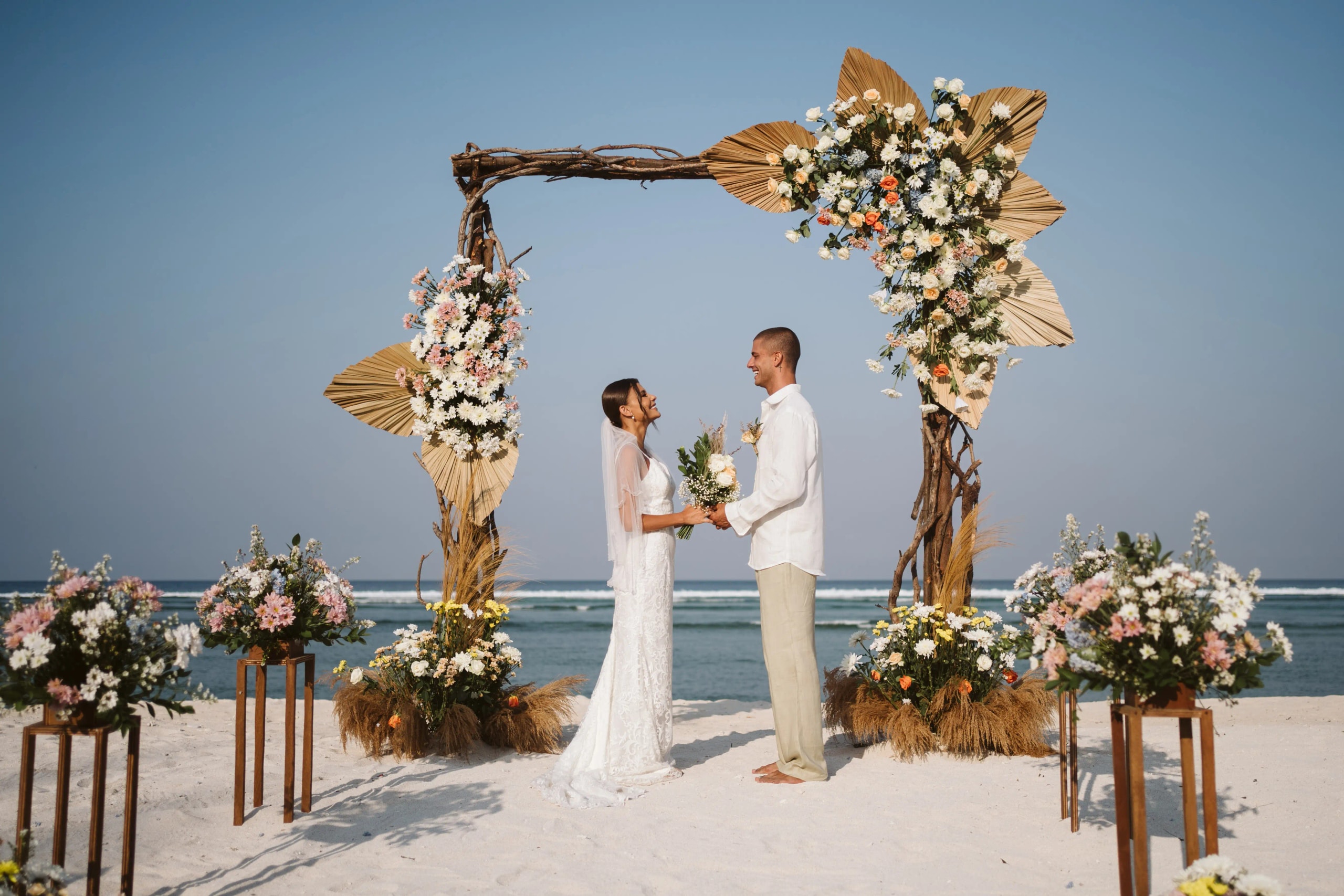 A bride and groom holding hands and smiling under a floral wedding arch decorated with dried palm leaves on a sandy beach.