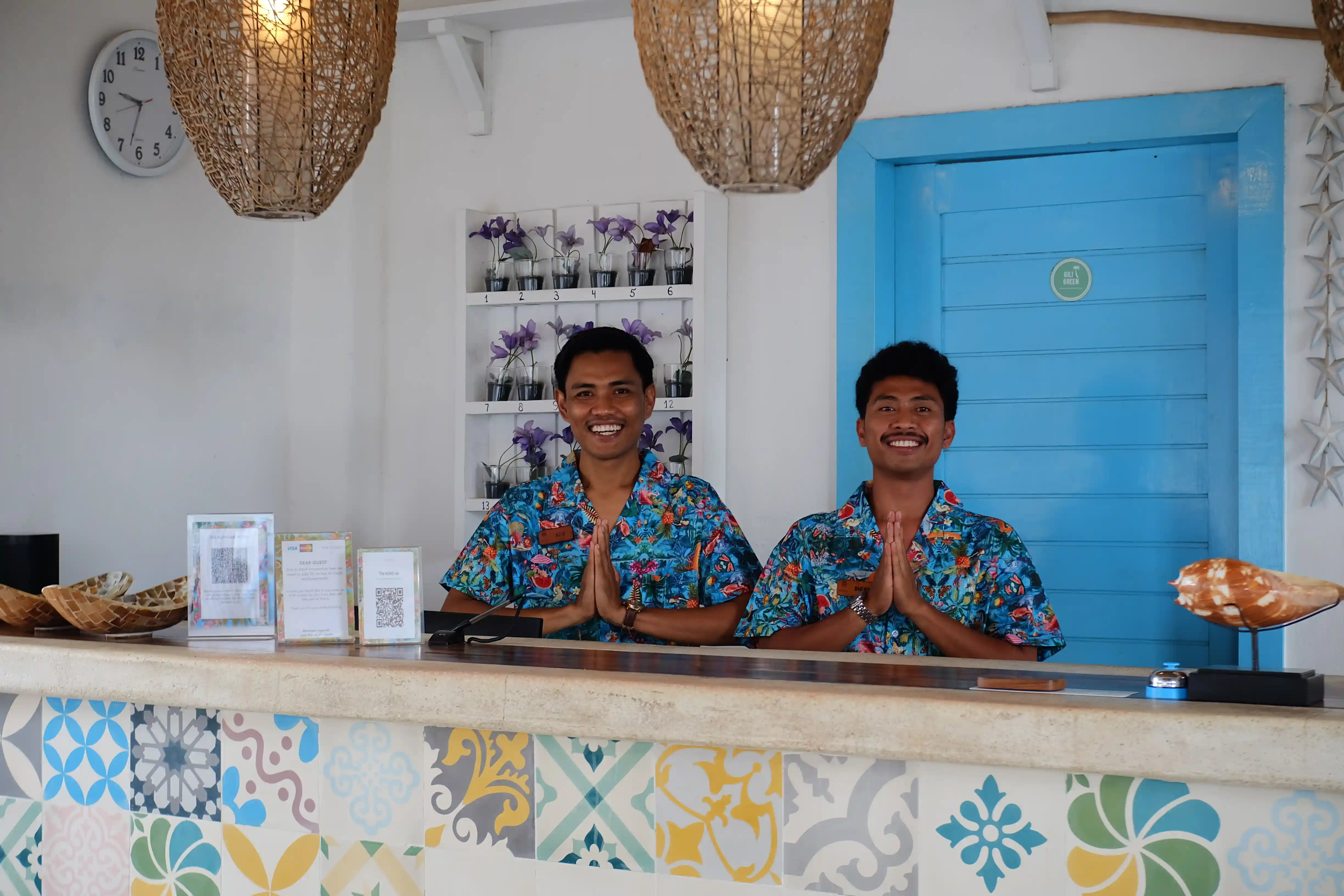 Deux réceptionnistes souriants en chemises tropicales faisant le geste de bienvenue derrière le comptoir.