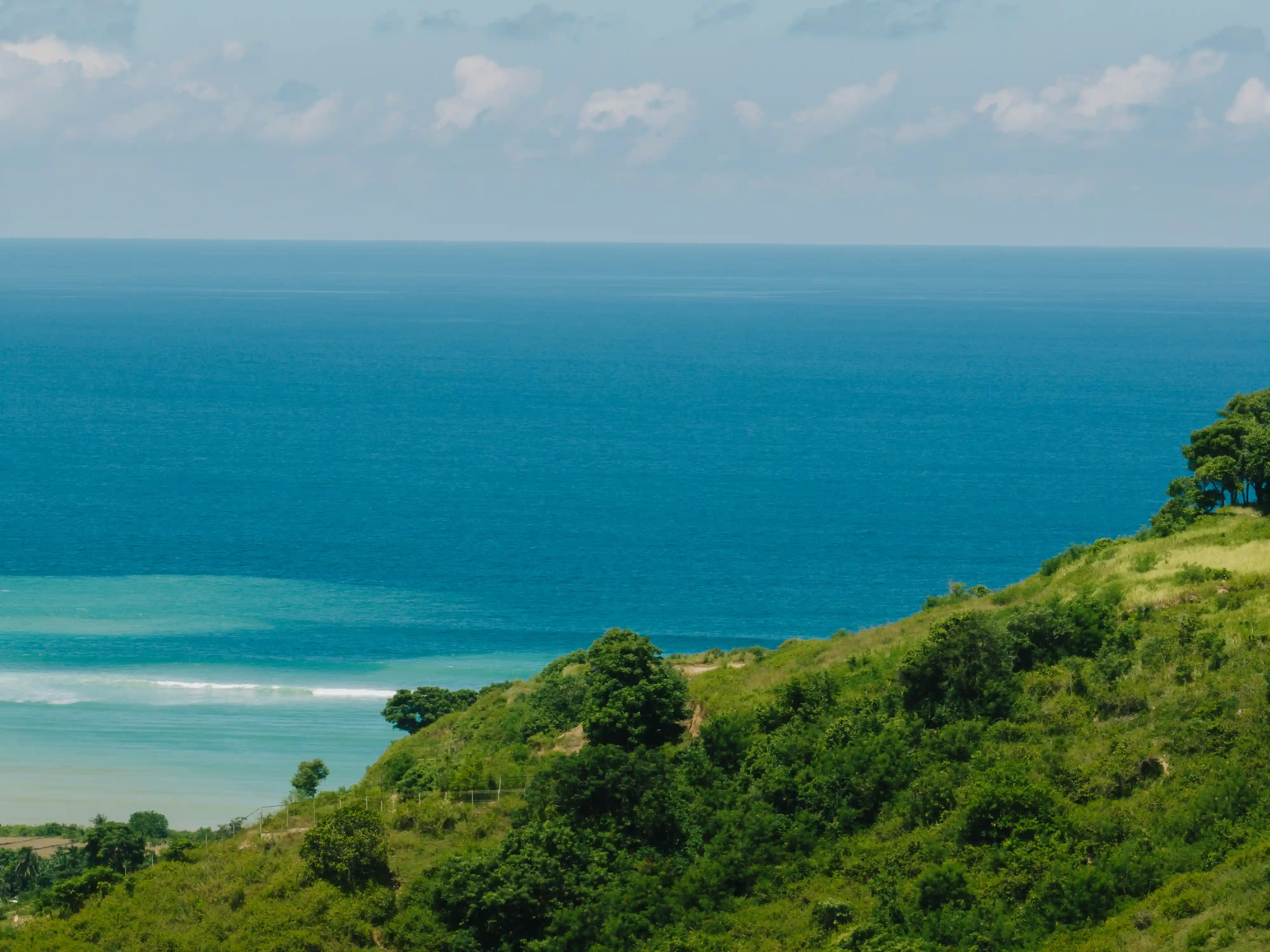 Lush green hillside sloping down toward vibrant turquoise ocean waves under a partly cloudy sky.