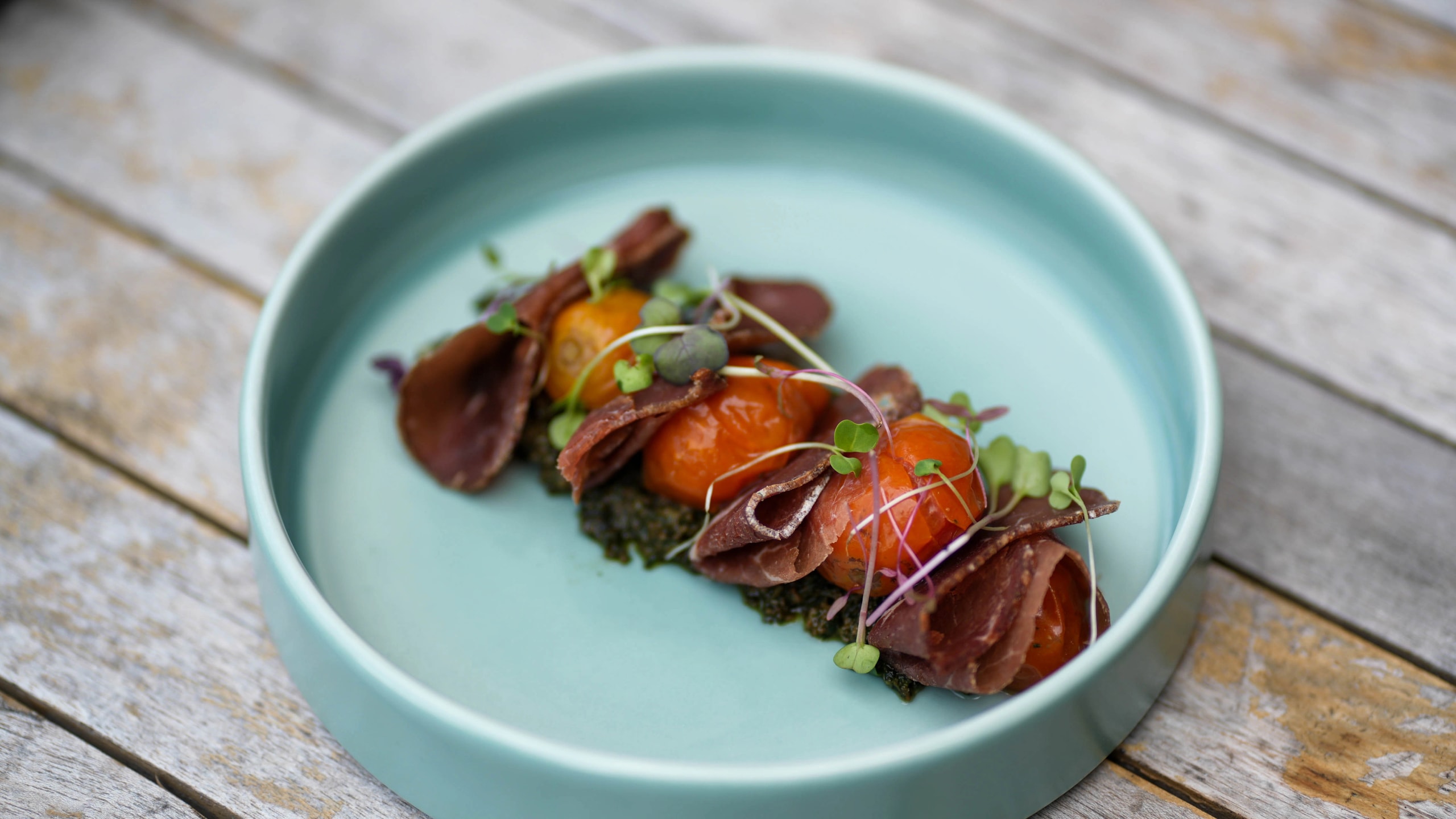 Cured meat rolled around roasted cherry tomatoes over green pesto in a light teal bowl.