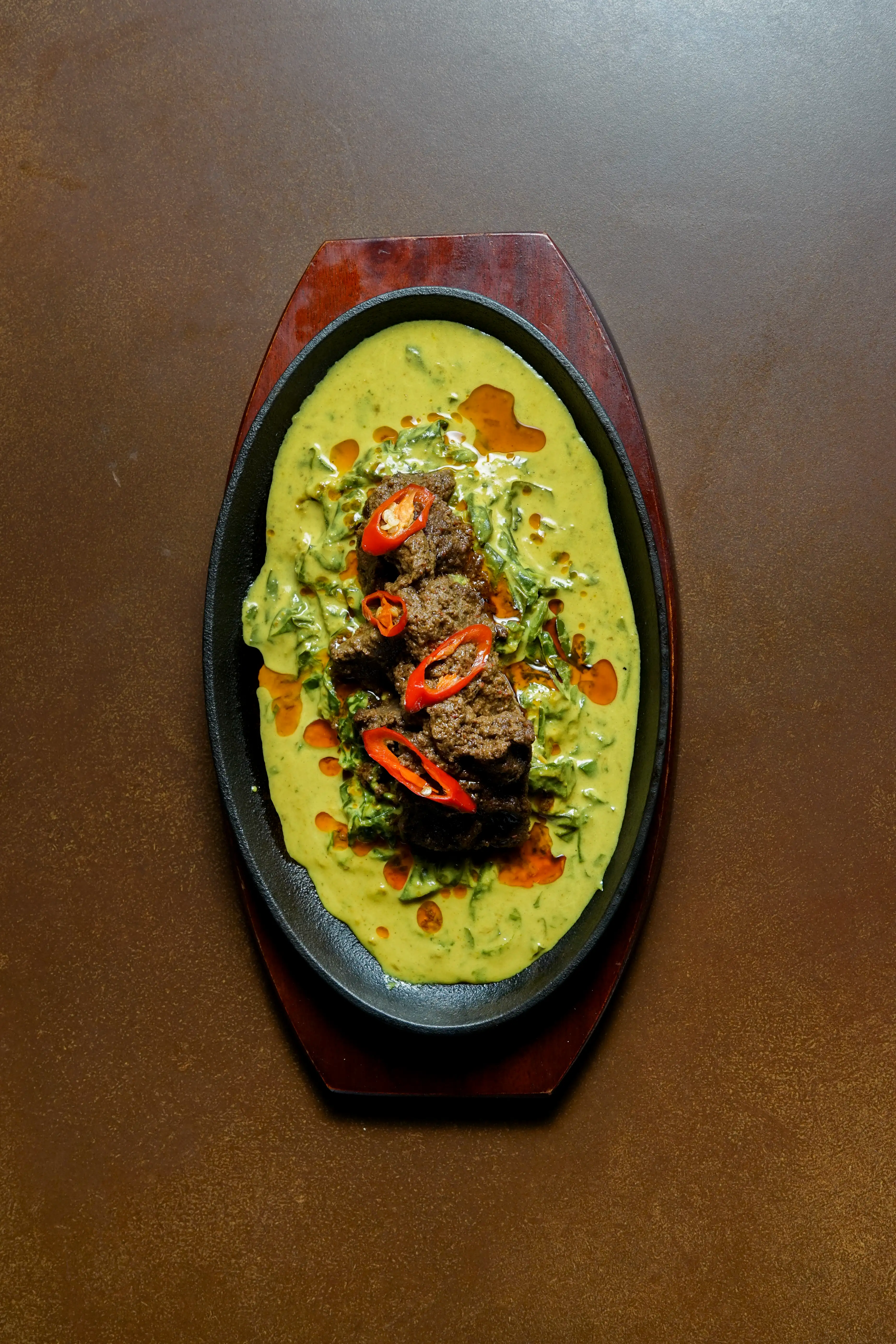 Top-down shot of beef rendang in a yellow curry base garnished with red chili on a black cast-iron platter.