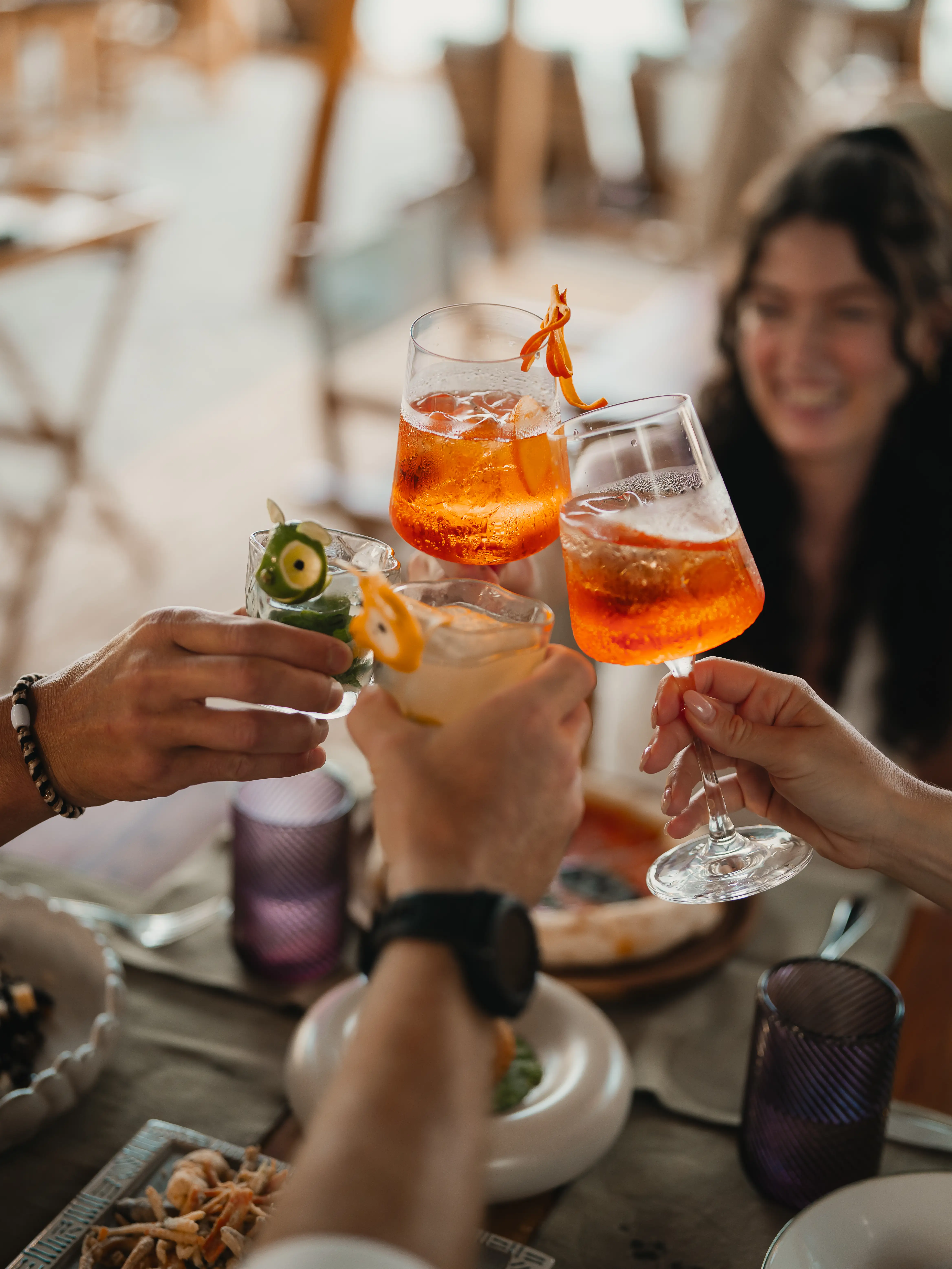 Friends clinking colorful cocktail glasses together in a celebratory toast at an outdoor table.