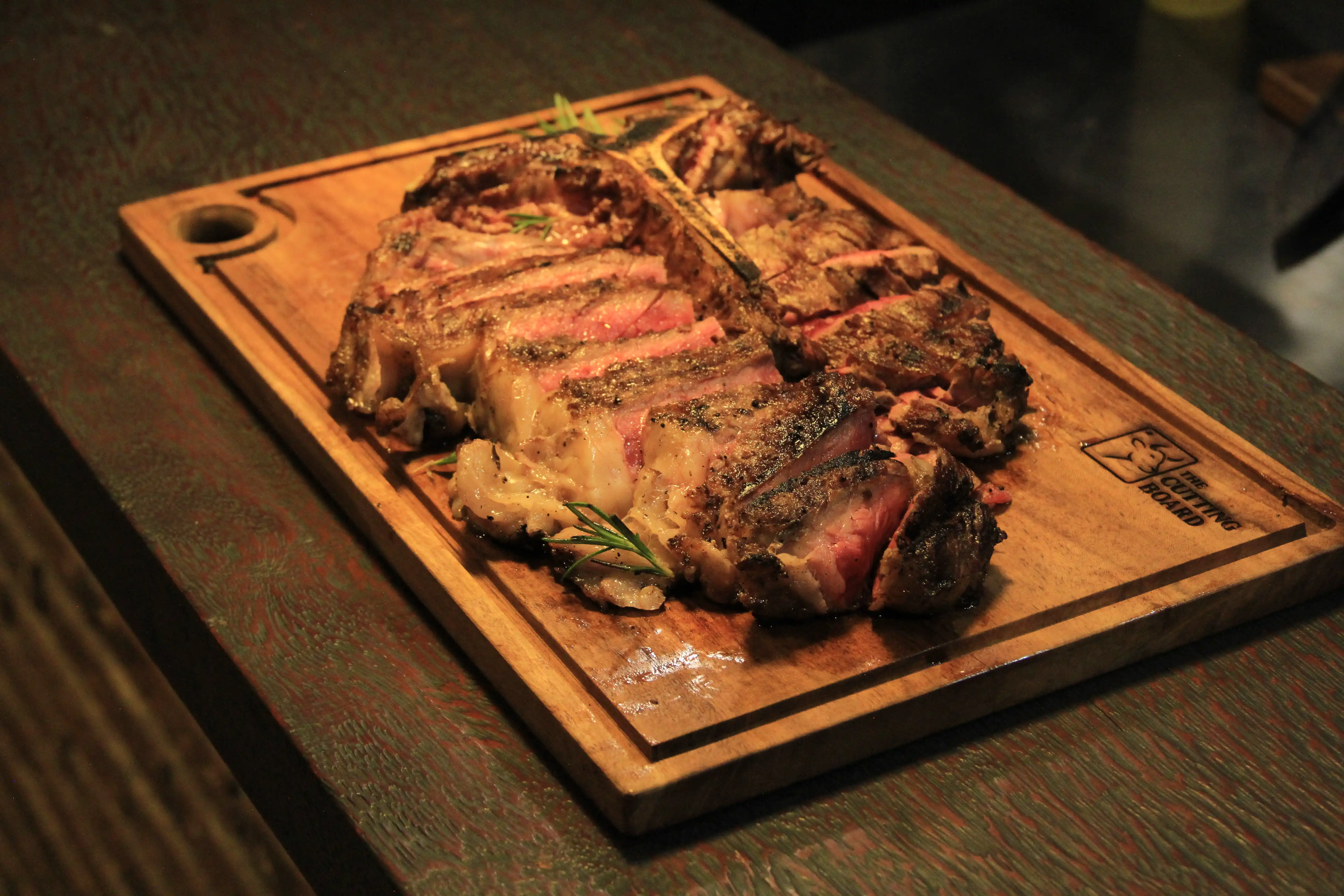 Sliced, grilled T-bone steak seasoned with rosemary, served on a branded wooden cutting board.