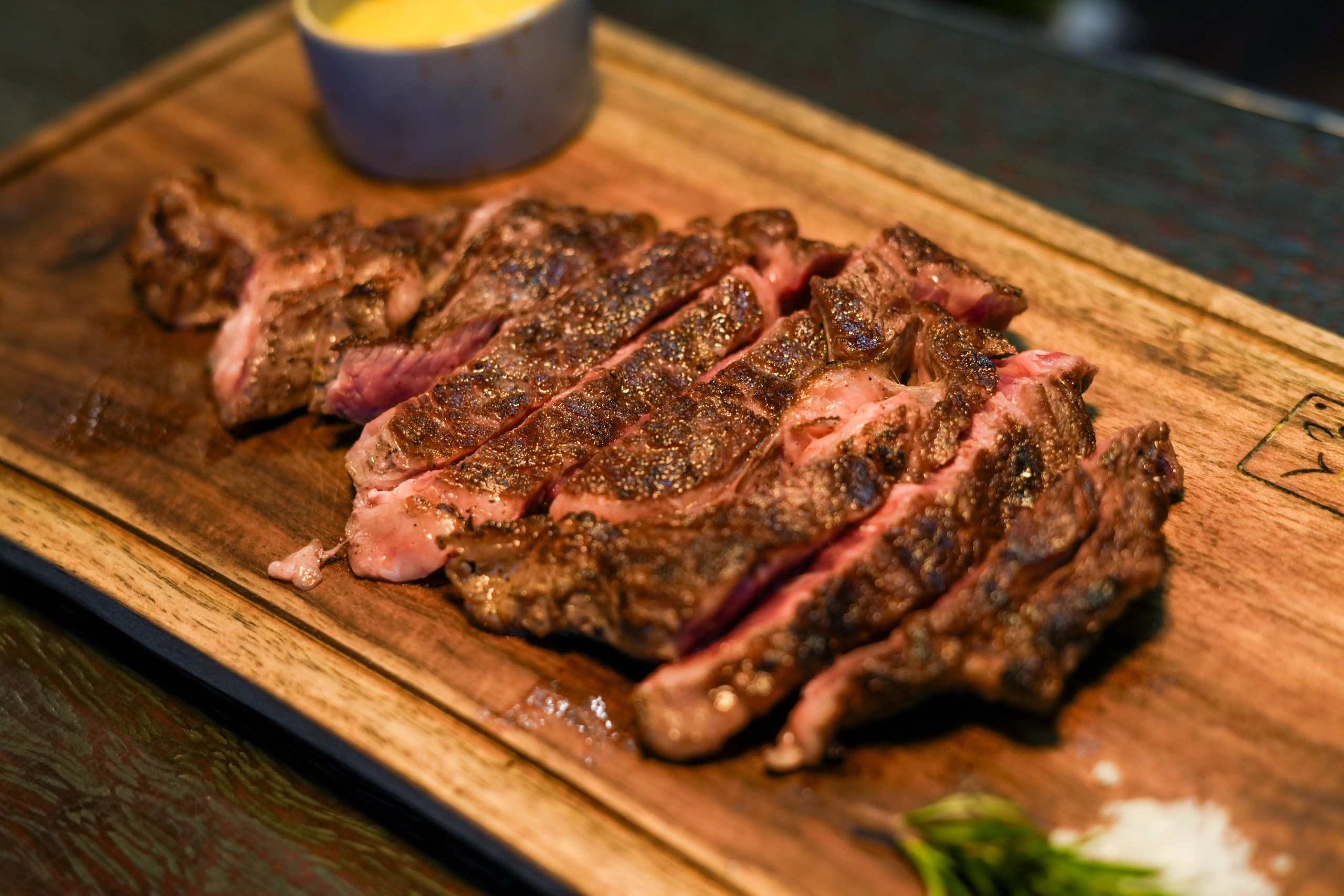 Sliced grilled steak served on a wooden board with sauce, sea salt, and herbs.