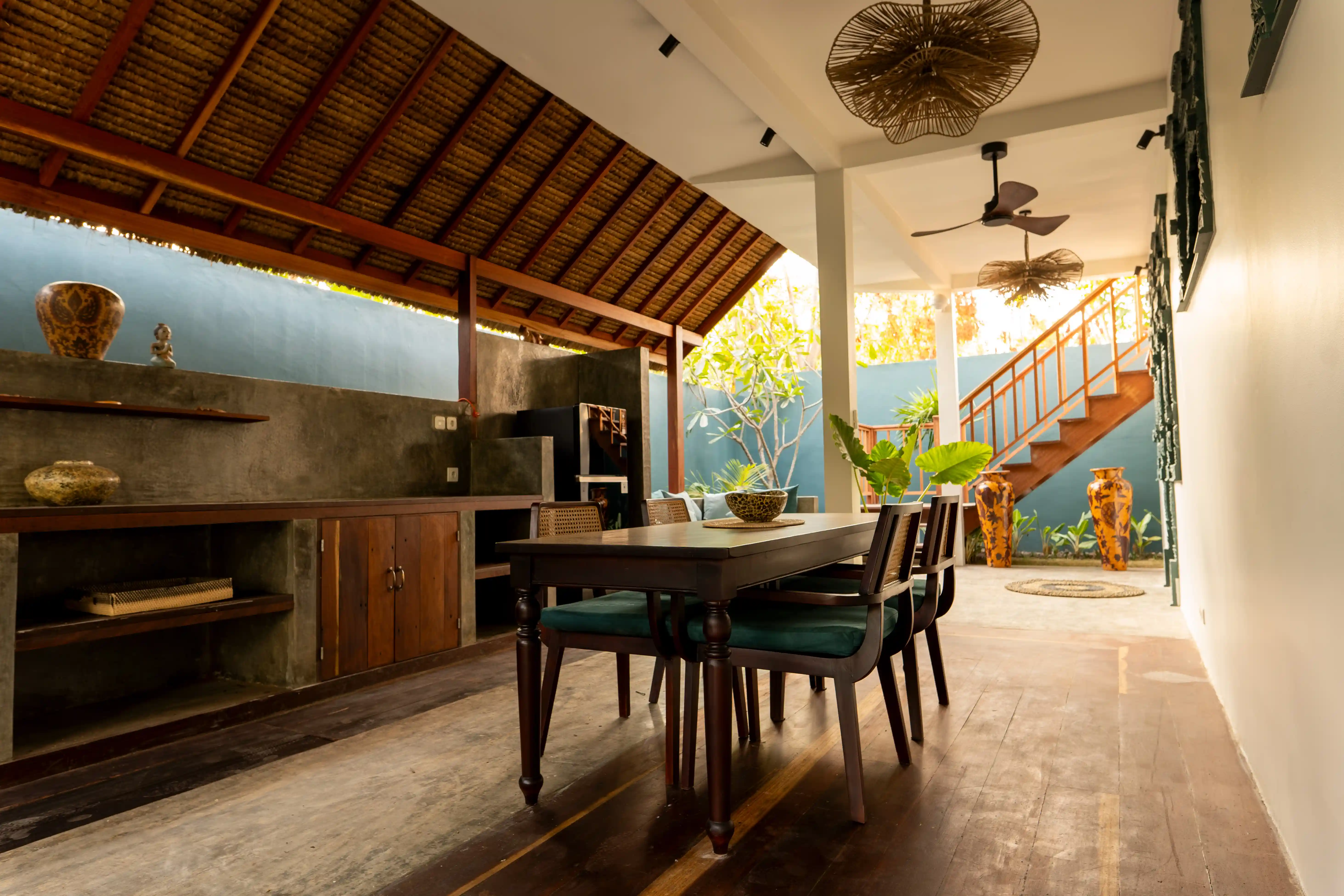 Open-air dining room and kitchen area featuring a dark wooden dining set, thatched roof, and a staircase in the background.