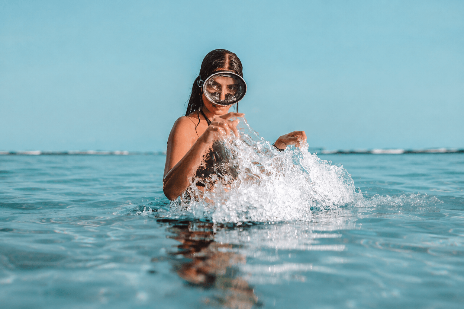 A woman wearing a diving mask surfacing in clear blue ocean water, splashing water with her hands.