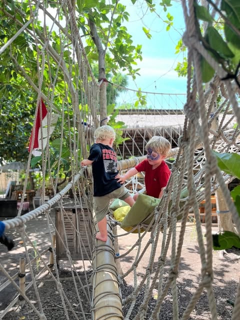 Two young children playing on an outdoor rope bridge structure in an adventure playground.