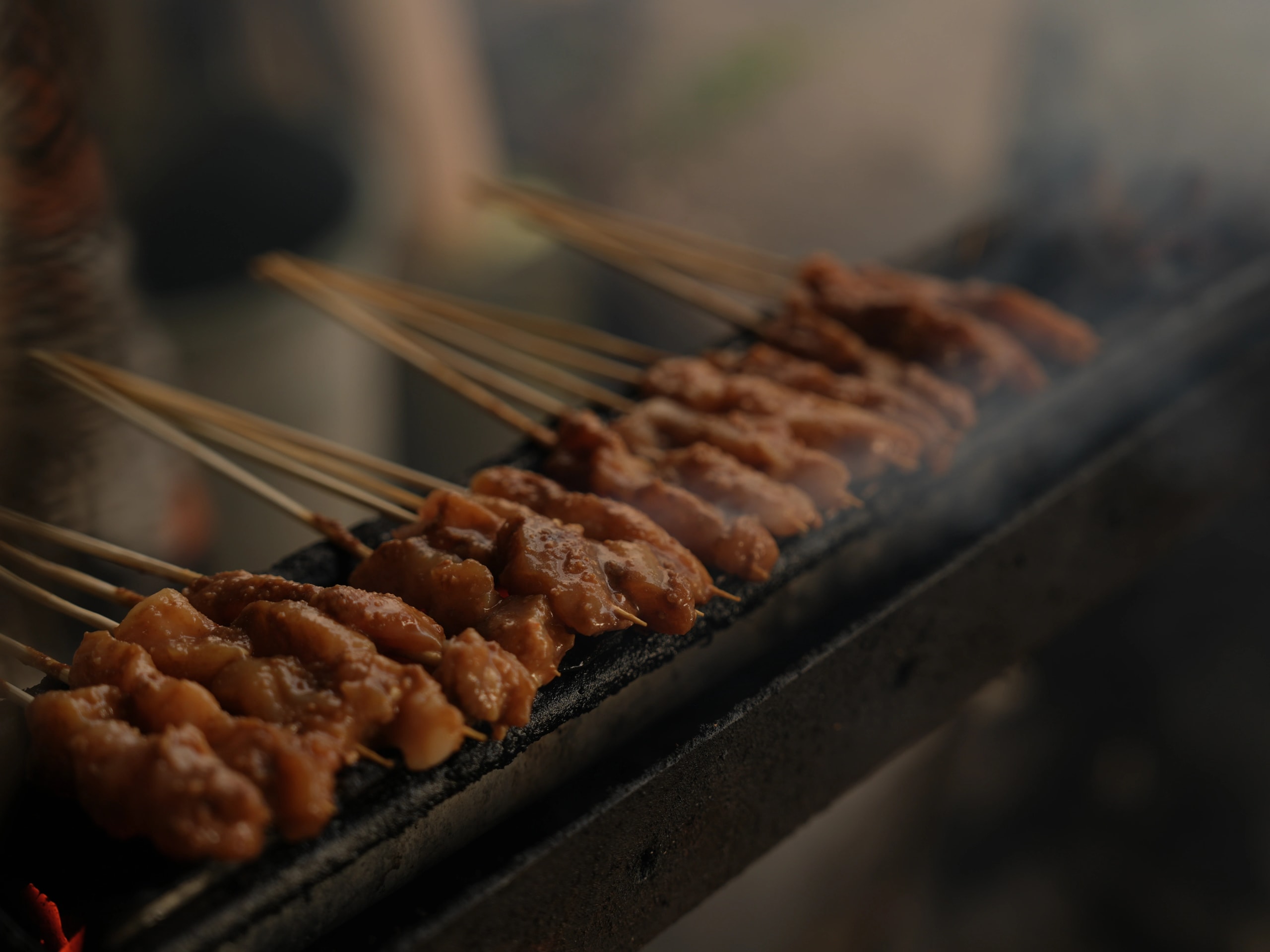 Close-up of chicken satay skewers grilling over charcoal with light smoke rising.