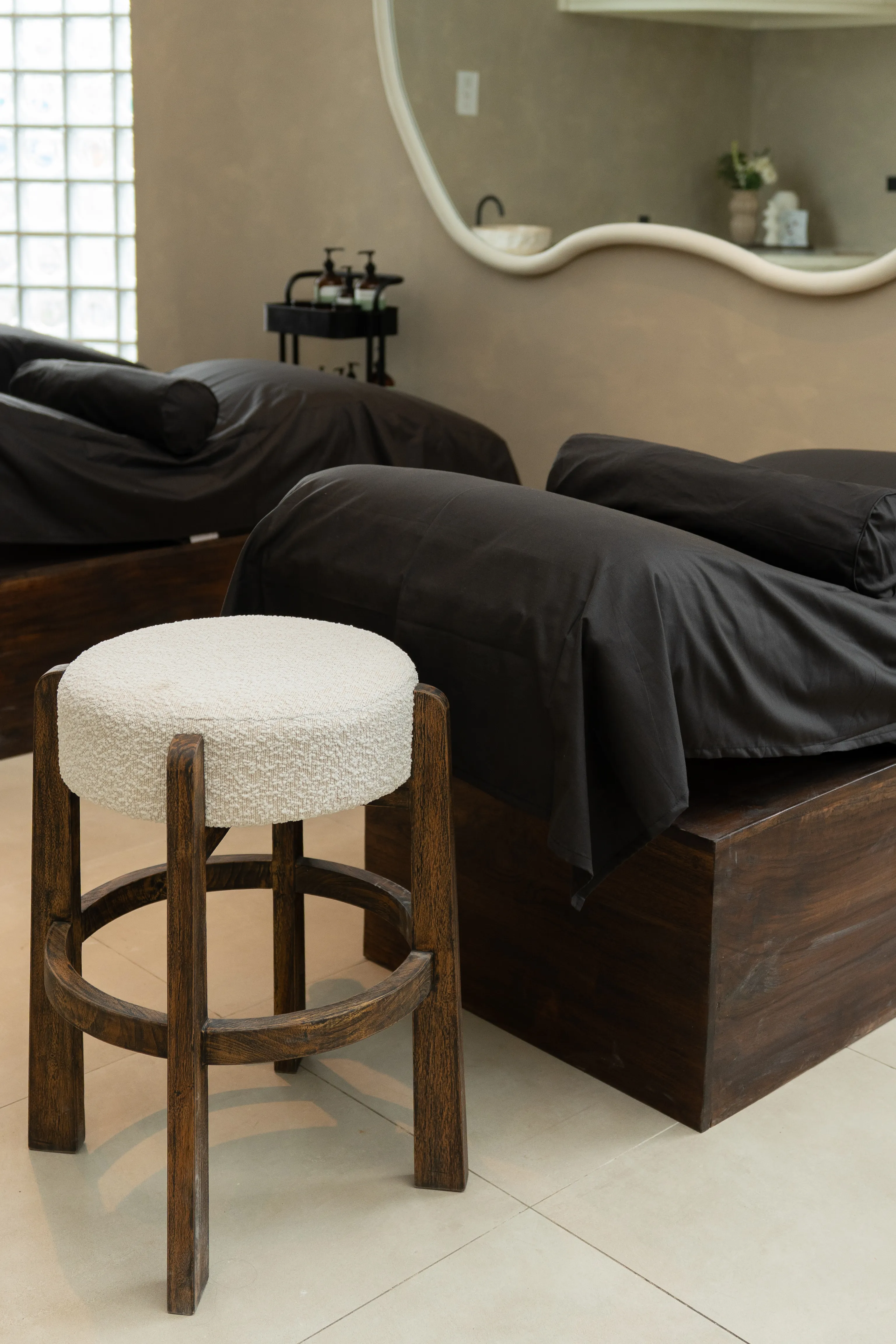 A modern spa treatment room featuring dark wooden massage beds dressed in black linens alongside a plush stool.