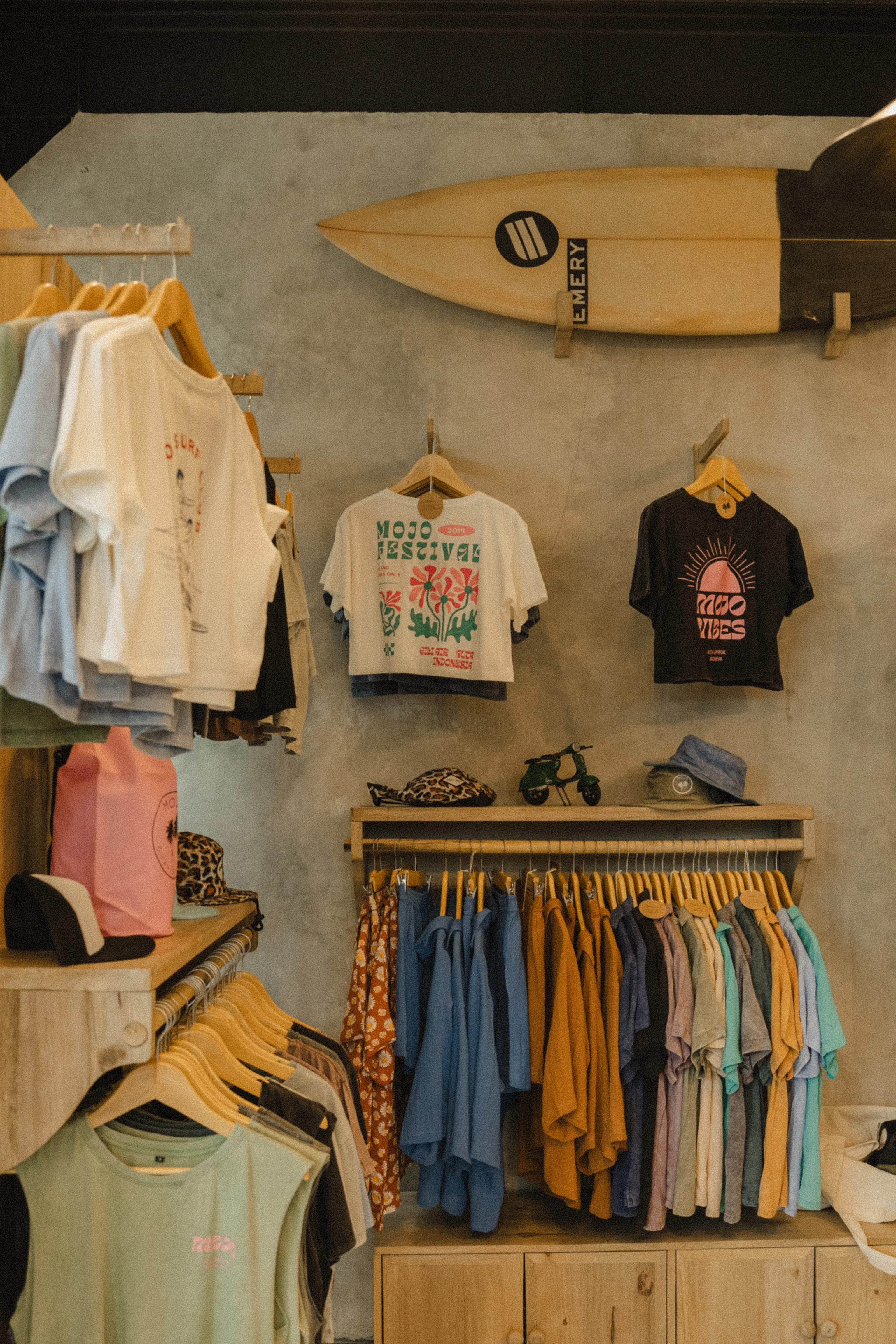 Interior of a surf shop featuring racks of colorful t-shirts and a surfboard mounted on a grey wall.