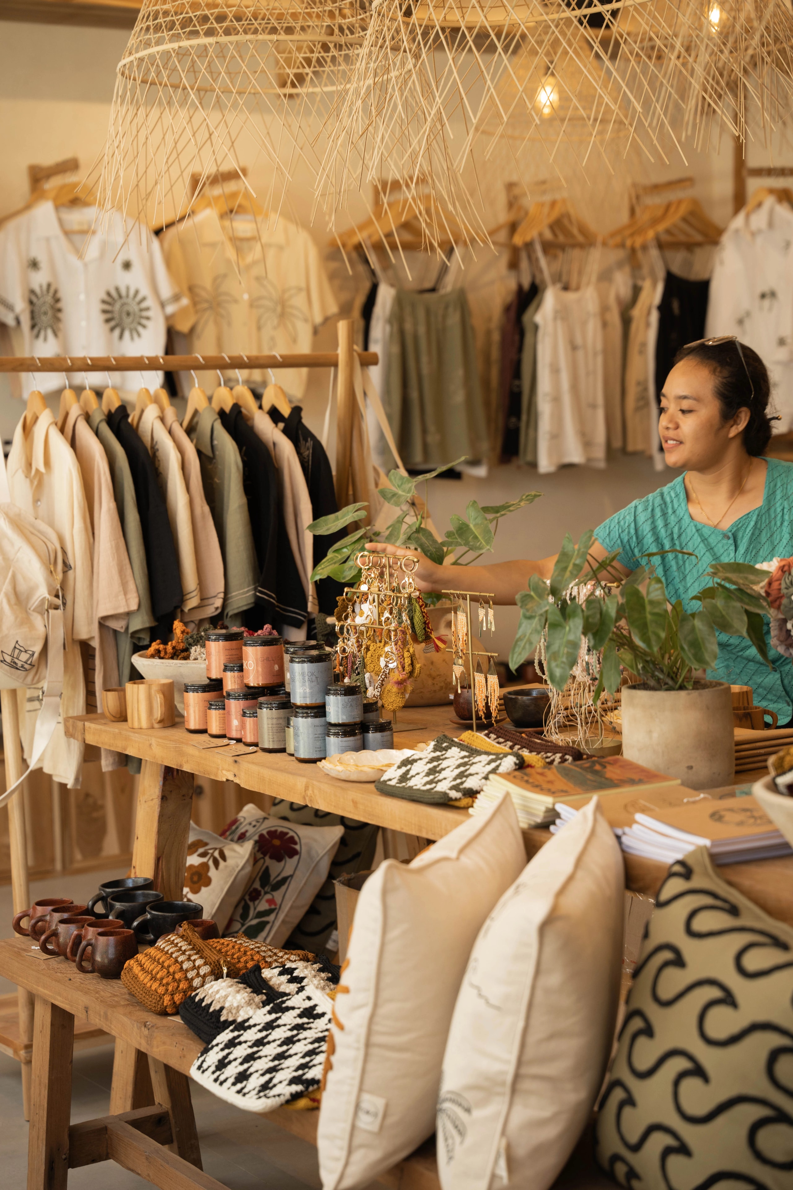 A woman arranging handmade accessories and candles on a wooden table inside a boutique clothing shop.
