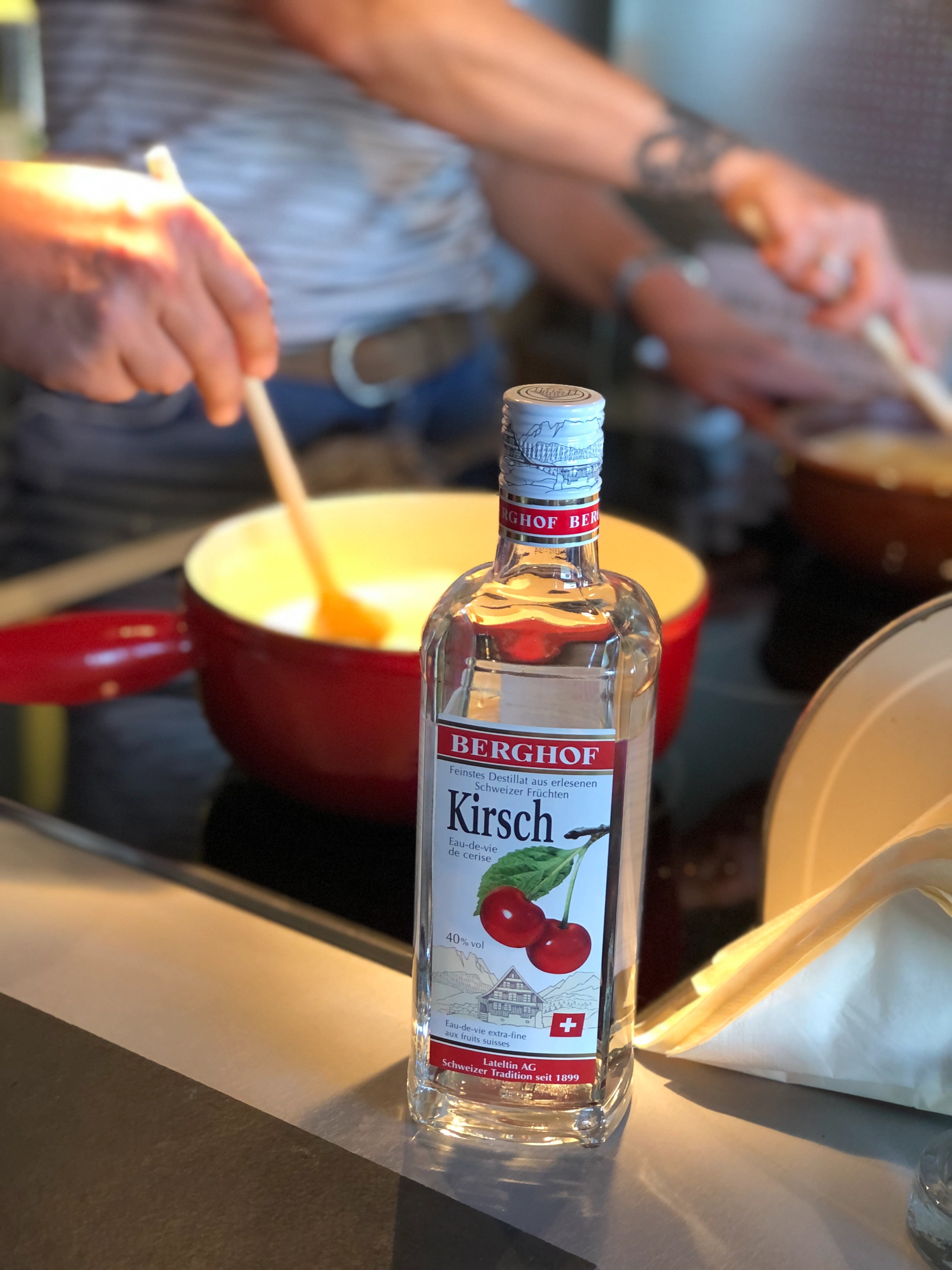 A bottle of Berghof Kirsch standing on a counter with someone stirring a fondue pot in the background.