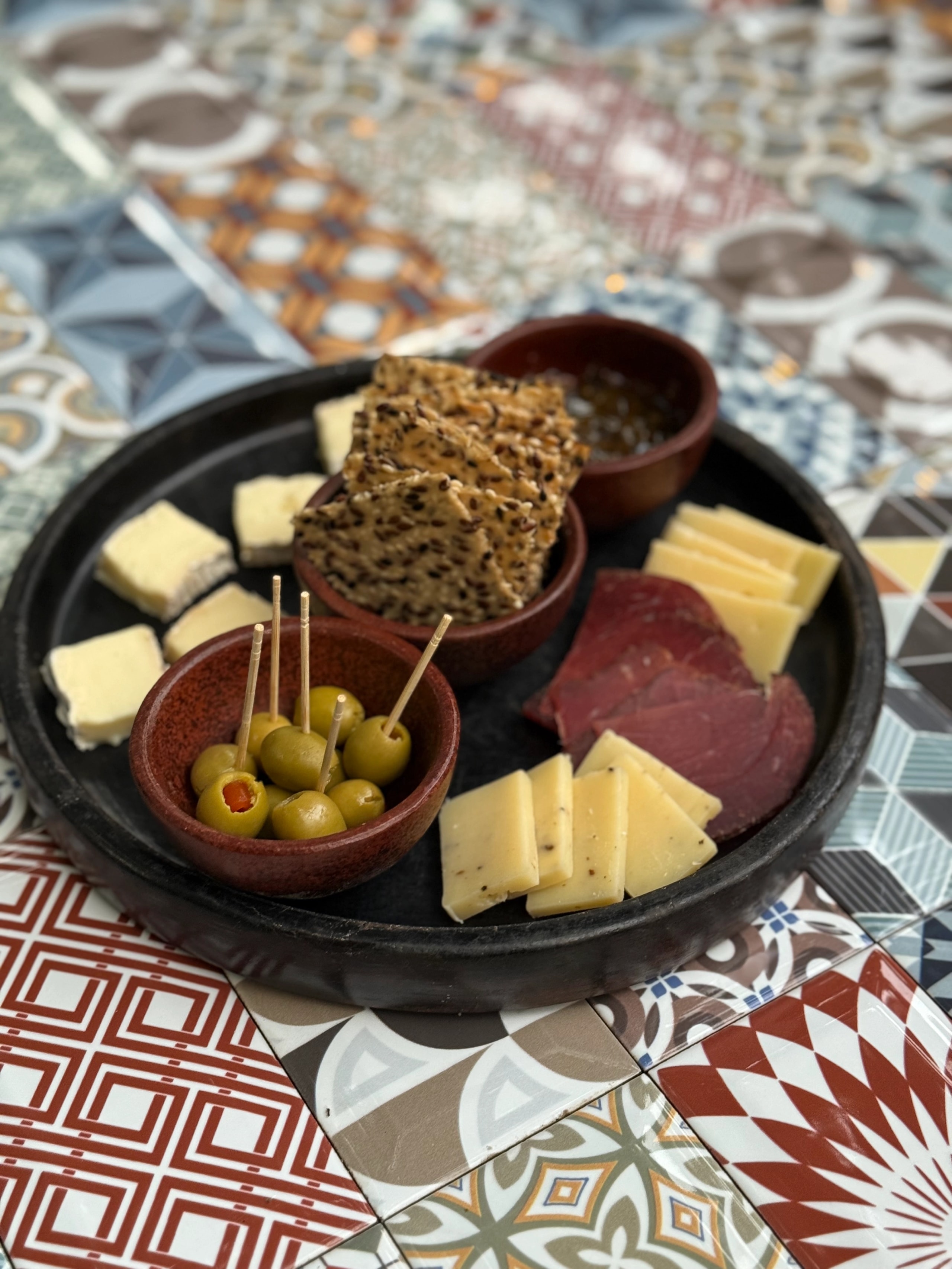 Charcuterie platter with assorted cheeses, cured meats, crackers, and green olives on a patterned tile table.