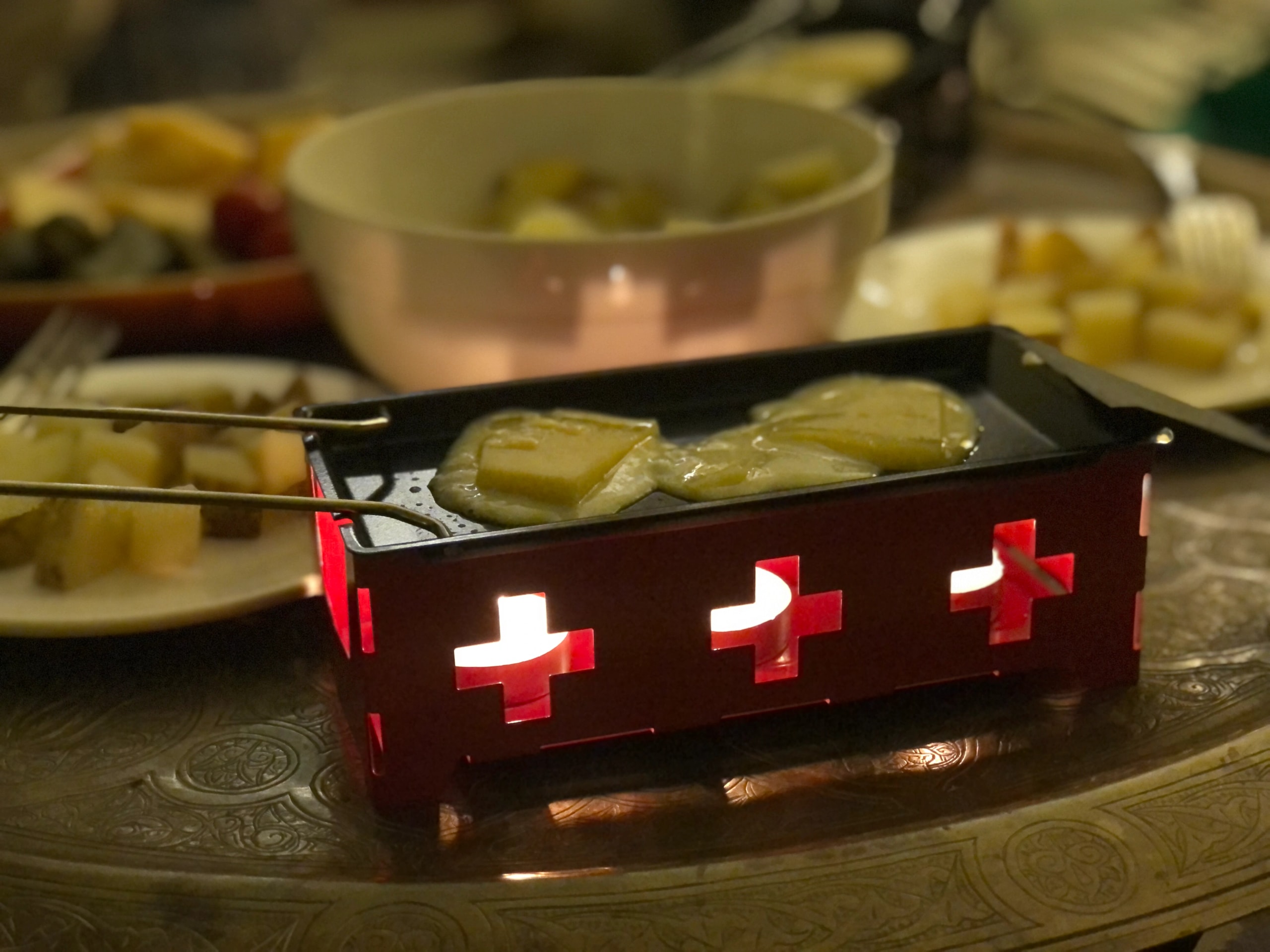 Tabletop raclette grill melting cheese slices over small candles inside a red box with Swiss cross cutouts.