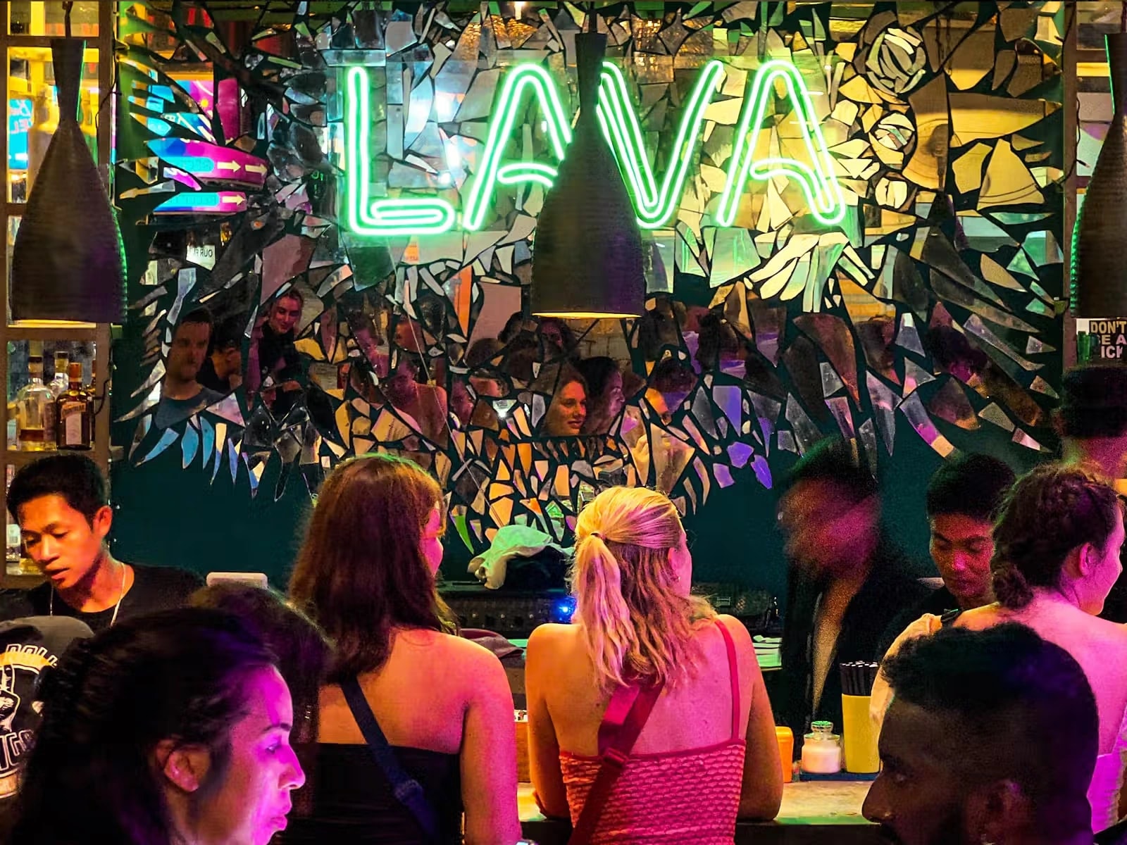 People gathered around a vibrant bar counter under a glowing green neon "LAVA" sign.