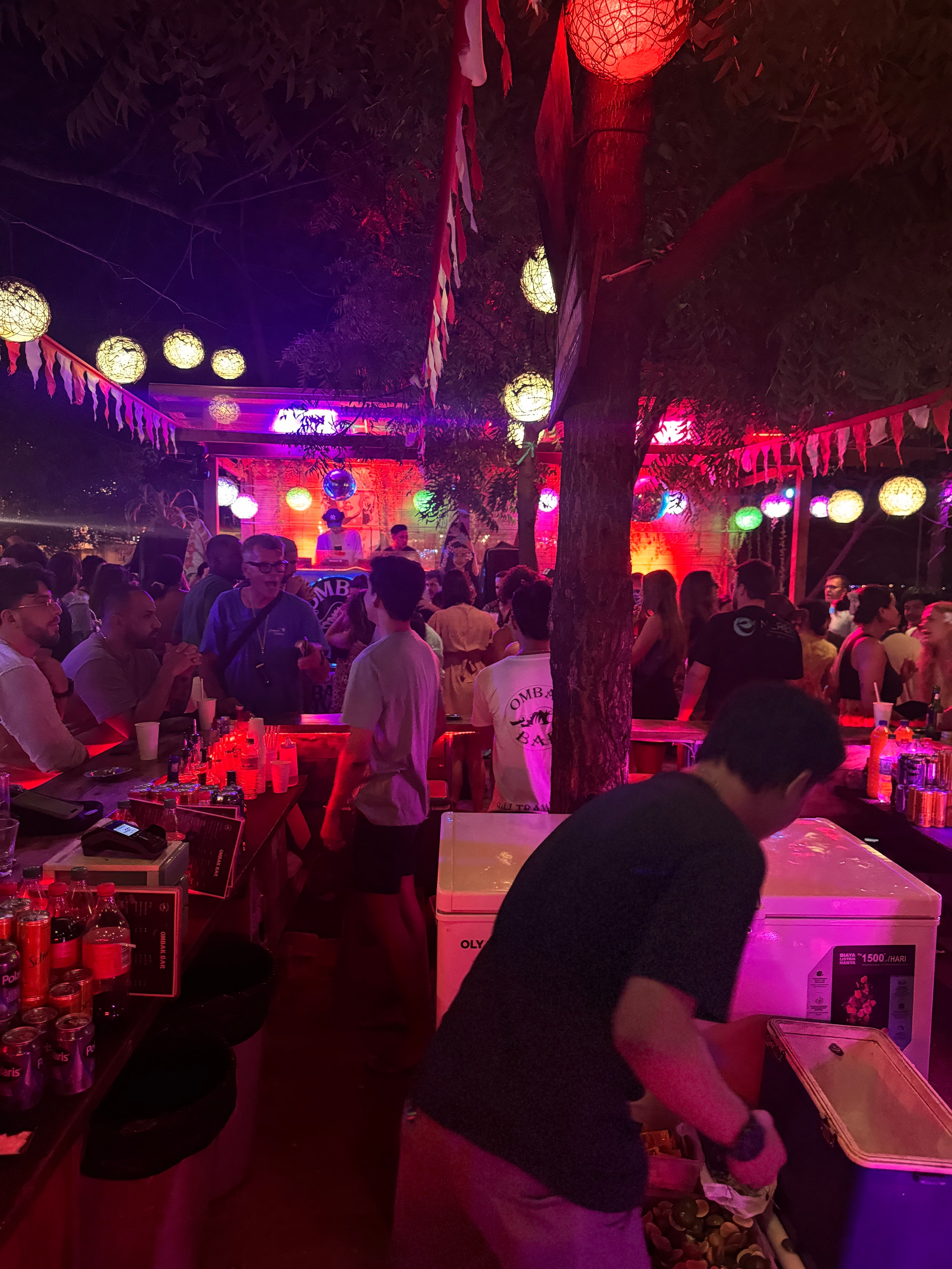 Crowded outdoor bar filled with partygoers dancing under glowing paper lanterns and red string lights.