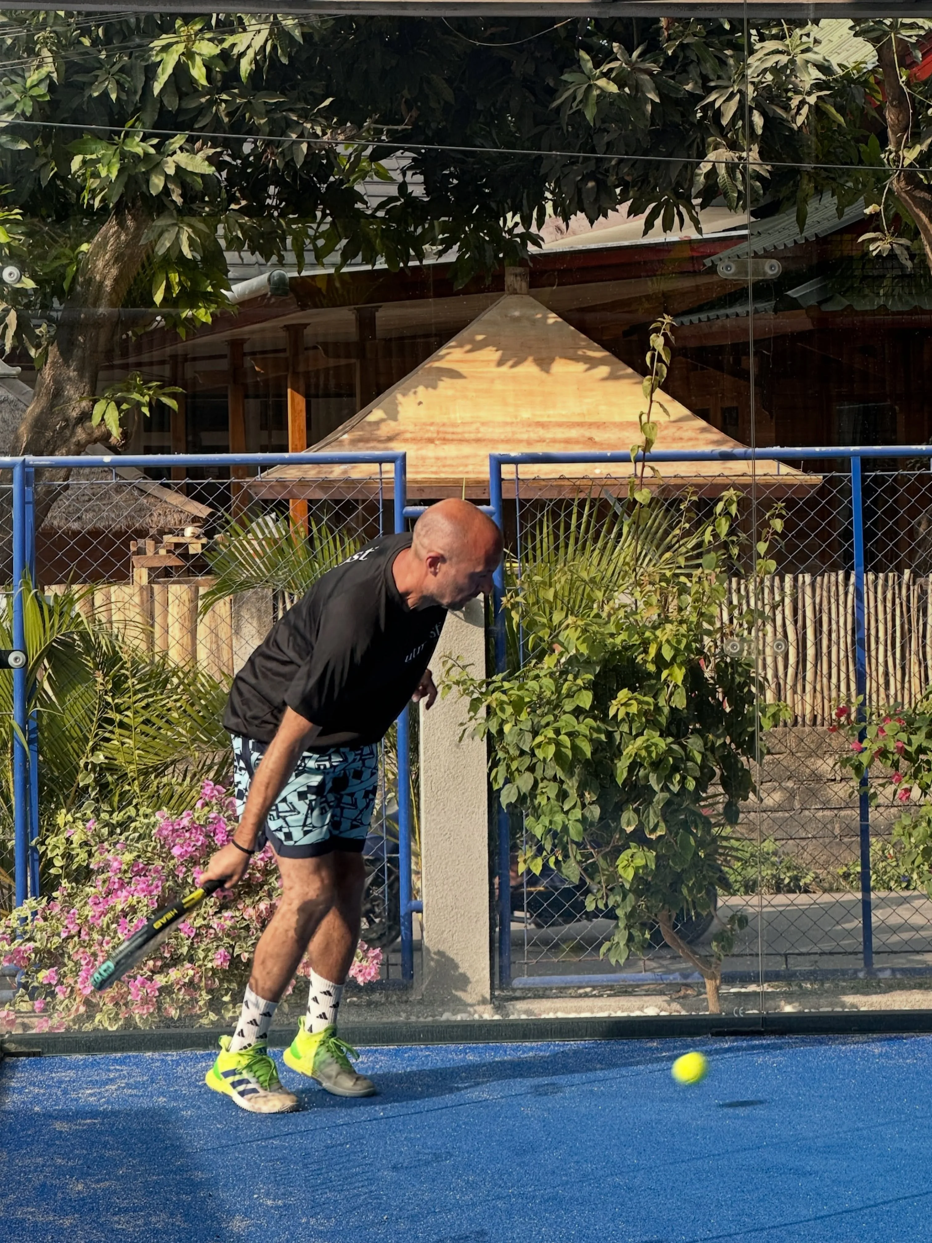 A man leaning forward on a blue padel court as a yellow ball bounces in front of him.