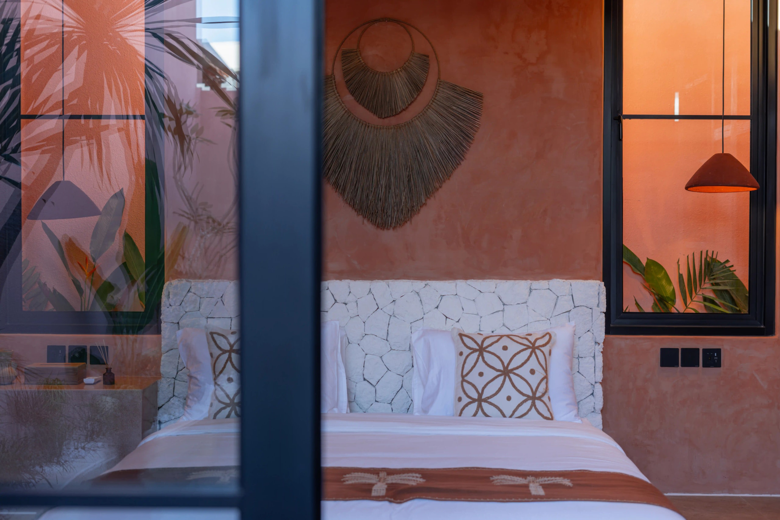 A bedroom view showing a bed with a white stone headboard and woven wall decor framed by dark-framed windows.