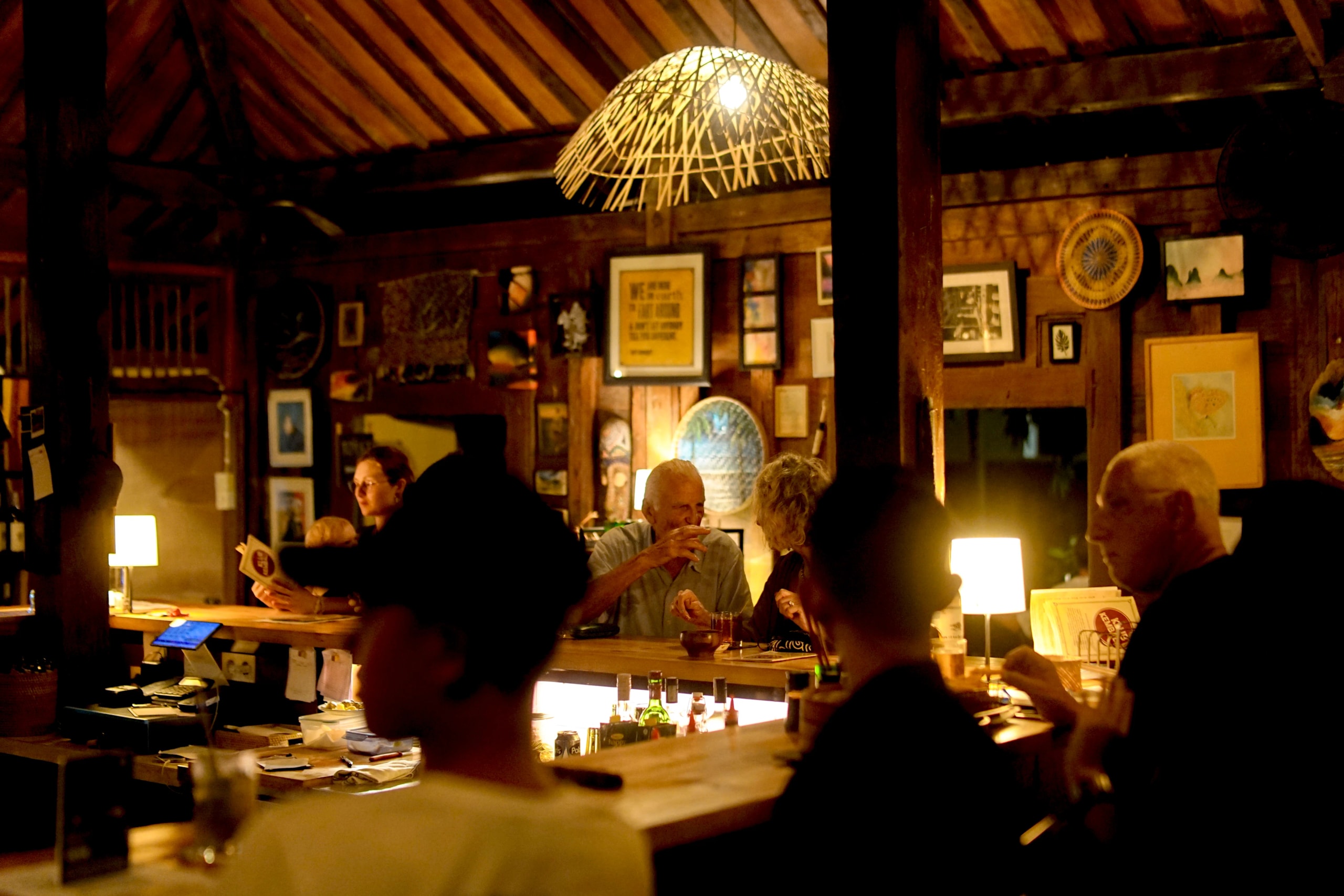Dimly lit rustic bar interior with patrons sitting at a wooden counter adorned with eclectic framed art.