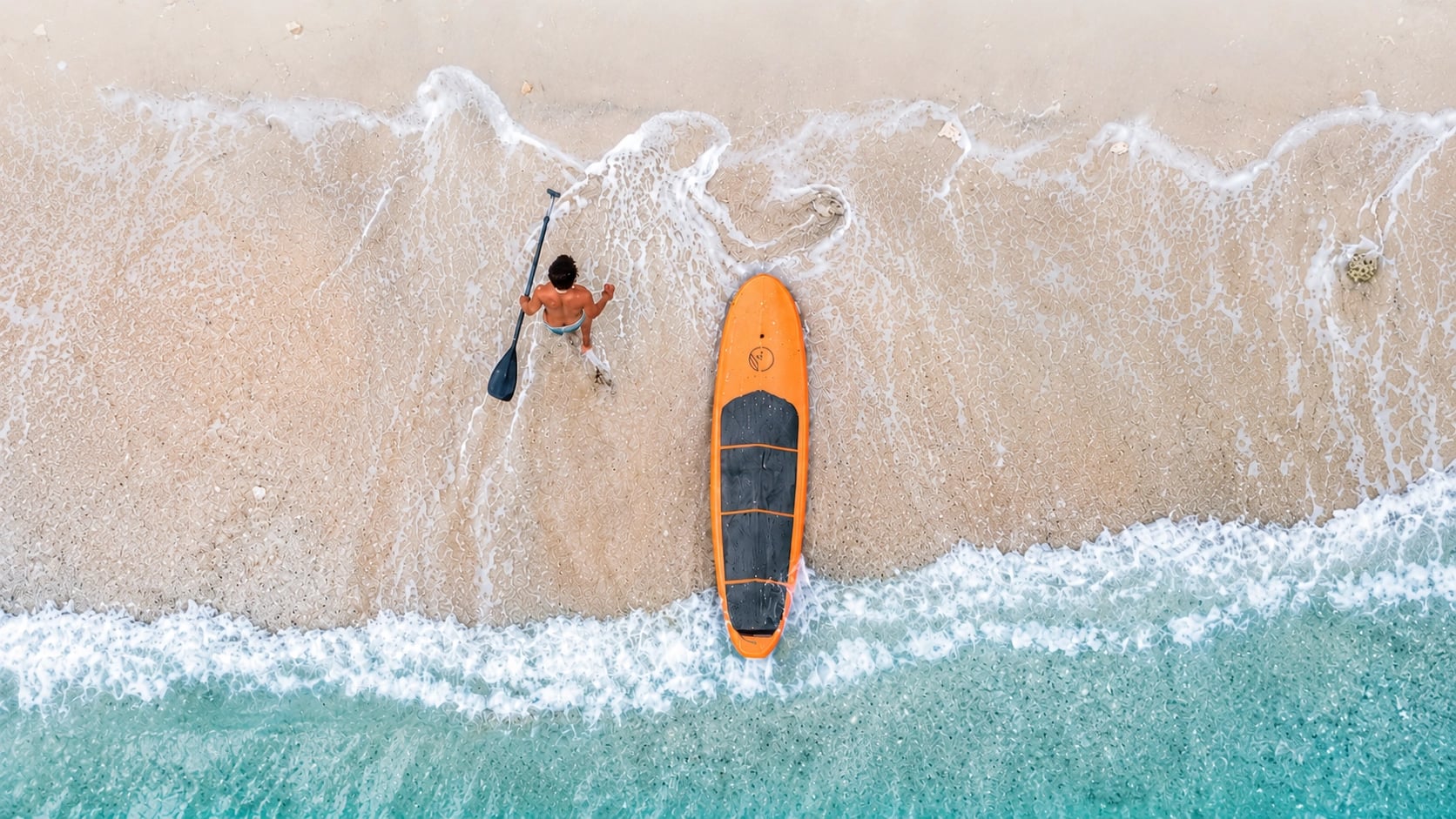 Aerial top-down view of a person holding a paddle next to an orange stand-up paddleboard on a sandy shoreline with gentle ocean waves.